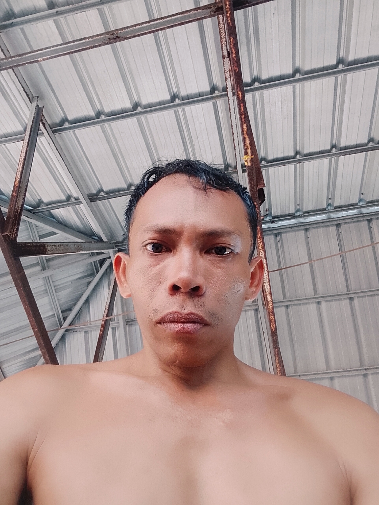 A shirtless man is visible in the foreground, shot from a low angle. His head and upper chest are in view. The background shows a metal roof structure with rusty support beams. The location is Bukit Ketok, Indonesia.