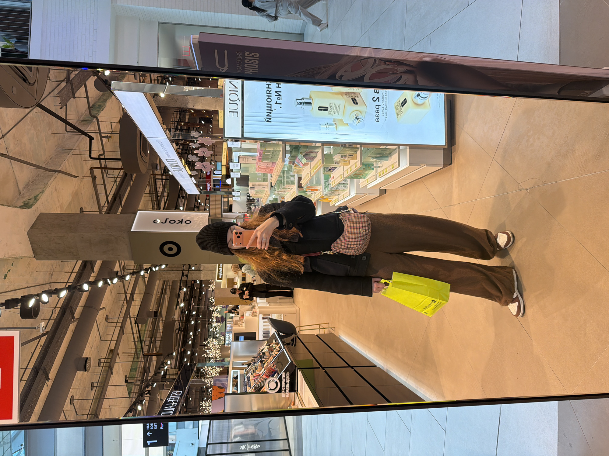 The image captures a mirror selfie taken by a young woman inside a modern shopping mall or large department store in Khimki, Russia. The woman, with long reddish-brown hair, is wearing a black beanie, a black jacket, and brown wide-leg pants with brown and white sneakers. She is holding a smartphone to capture her reflection and carries a bright yellow shopping bag in her left hand, along with a small plaid-patterned shoulder bag.
The setting is a well-lit indoor retail space, characterized by light-colored tiled flooring and an industrial-style ceiling featuring exposed pipes and track lighting. Numerous retail displays and advertising billboards are visible in the reflection, showcasing various products, including cosmetics and perfumes (e.g., Clinique). Other shoppers are faintly visible in the background, suggesting a busy environment.
Visible text and brands include "Joko," "VIONE," "PHOSIS" (as part of "КОЛЛЕКЦИЯ PHOSIS" - "PHOSIS COLLECTION"), and "CLINIQUE." Other Russian text such as "ДЛЯ ОТКРЫТИЯ НАЖАТЬ ЗДЕСЬ" ("TO OPEN CLICK HERE"), and various mirrored/upside-down words like "NNWIRH," "ГИ Н," "ERLEEFYOD," and parts of promotional material like "АЖOX RAHHEНЖАЛА" are present. A directional sign indicates "↑ MAA BOS 12ST." The overall impression is a casual shopping excursion.