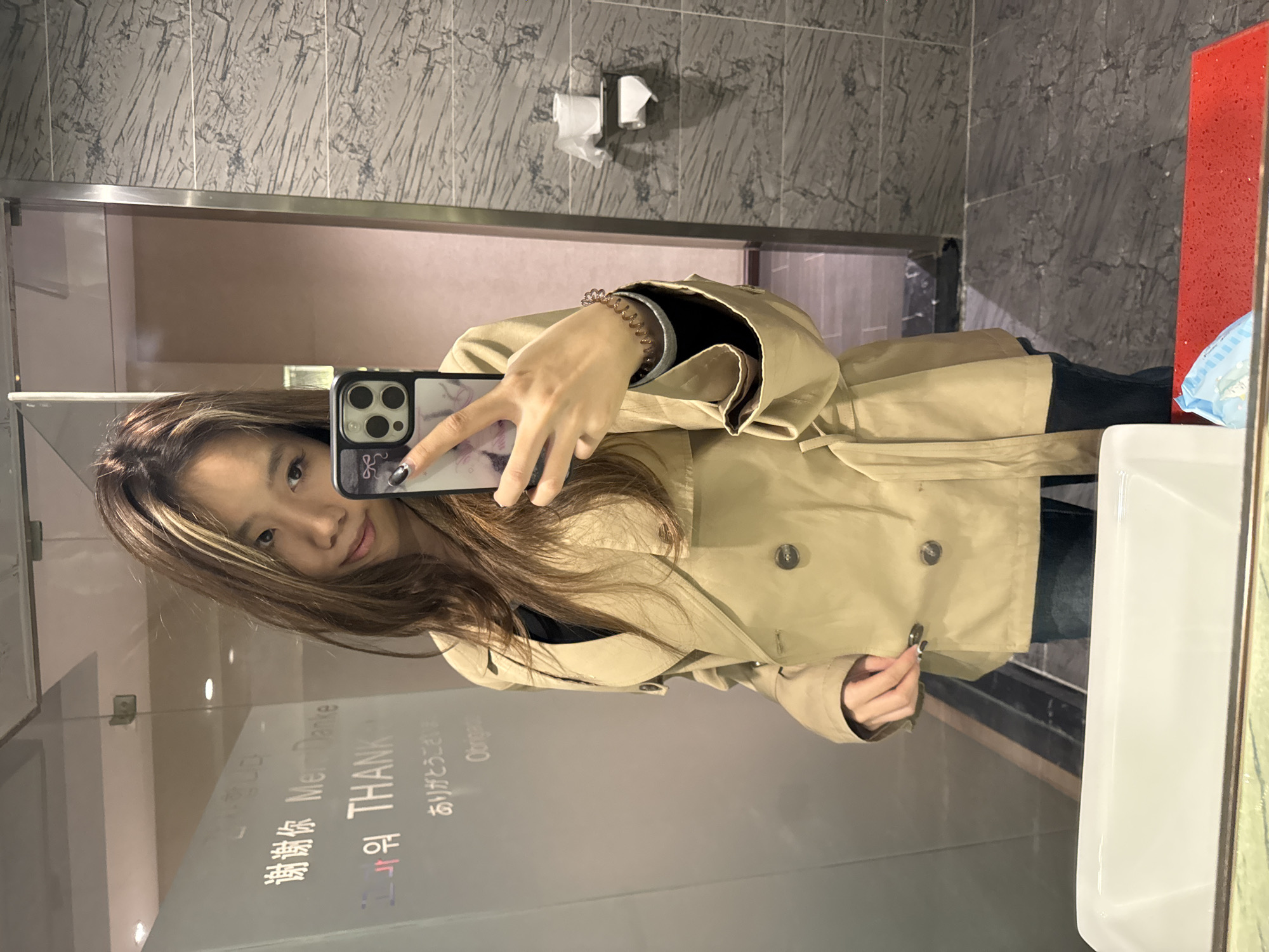 A young East Asian woman is captured taking a mirror selfie in what appears to be a public restroom or washroom. She has long, dark brown hair with blonde or light brown highlights and is wearing a beige or khaki double-breasted trench coat over a dark top, paired with dark trousers or jeans. She holds a black smartphone, likely an iPhone due to its distinctive camera array, in her left hand, which has a patterned case. She looks directly at her phone, with a neutral to slightly smiling expression. A spiral hair tie or bracelet is visible on her right wrist.
The setting features grey, marble-patterned tiled walls. Behind her, two rolls of toilet paper are on a wall-mounted dispenser. To her right, a white ceramic sink and a reddish-orange, textured panel are visible, along with a blue and white patterned object, possibly a small bag, resting on the counter. The lighting is artificial, suggesting an indoor environment.
Reflected on the mirror are various translations of "Thank You," including "谢谢你" (Chinese), "Mer Danke" (resembling German "Danke"), "THANK" (English), "고마워" (Korean), and "ありがとうごさいます" (Japanese). These multilingual greetings indicate a public space catering to diverse visitors, located in Ganzhou, China, as per context.
