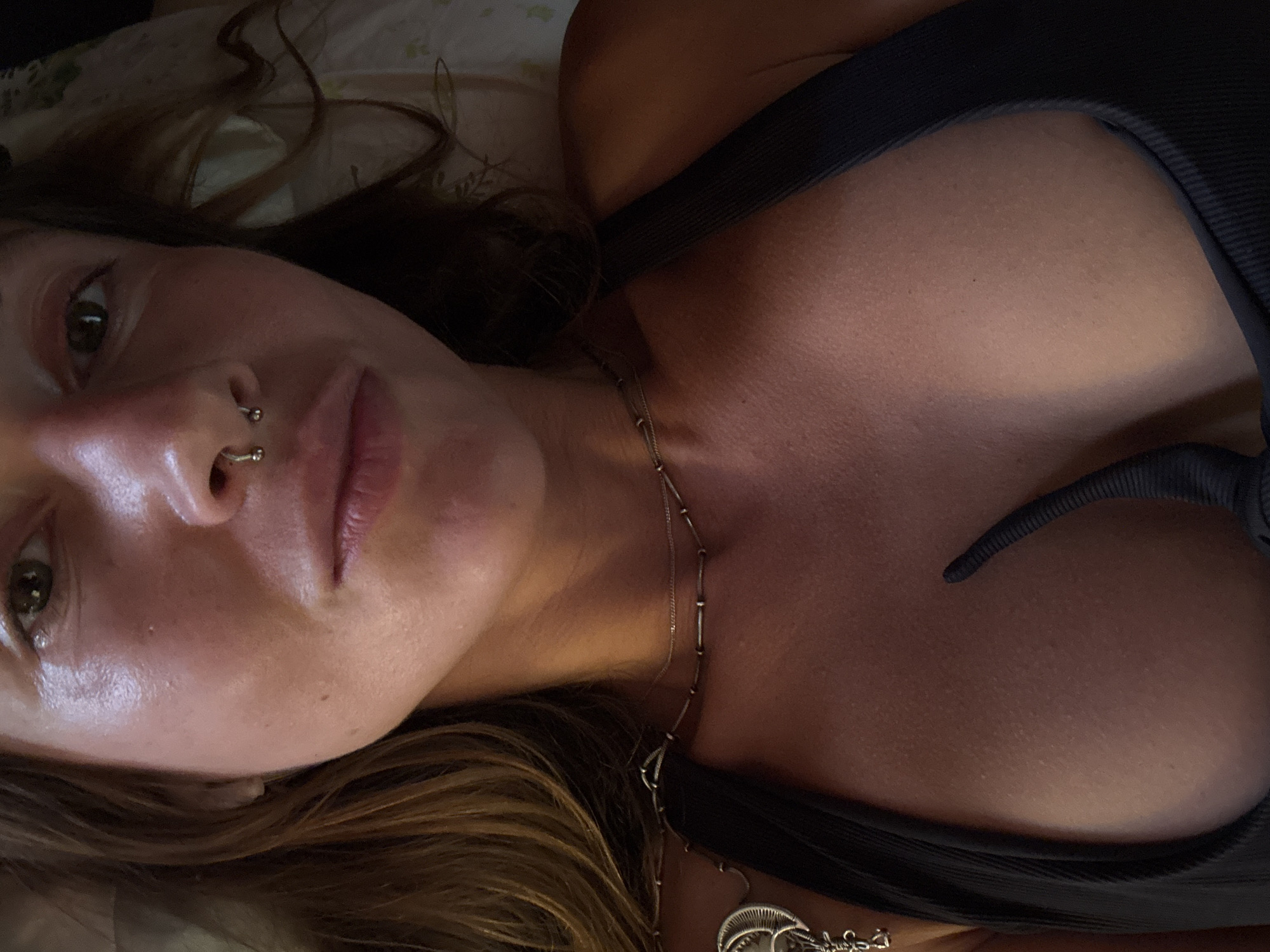 A close-up, chest-up shot shows a woman with brown hair and a septum piercing. She is wearing a black top and a necklace. The lighting is warm and highlights her skin. The background is blurred and appears to be a bed with patterned sheets.