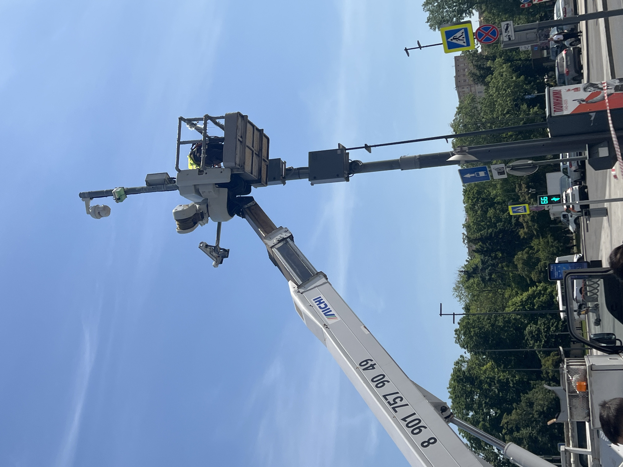 An outdoor urban scene in Moscow, Russia, captured on a clear, sunny day. The primary focus is a worker elevated in the basket of a white "MICHI" brand cherry picker (articulated boom lift) while performing work on a dark grey street pole. The cherry picker's boom prominently displays the Russian phone number "8 901 757 90 49". The worker, partially visible within the metal basket, wears a yellow high-visibility vest and a hard hat.
The street pole supports various components: two surveillance cameras (one dome-shaped, one cylindrical), and two grey rectangular electrical enclosures mounted at different heights. Below these, a pedestrian traffic light displays the countdown "66". Multiple Russian standard traffic signs are attached to the pole and adjacent structures, including a blue square "pedestrian crossing" sign, a blue square "one-way street" or "bus lane" sign with an arrow and a bus icon, a white rectangular sign with a black camera icon (indicating surveillance), a red circular "no stopping/standing" sign, and a white rectangular "tow away zone" sign.
In the background, lush green trees, indicative of an urban park (Alexander Garden), line the left side of the frame. Through these trees, a large, multi-story building with a light-colored, classical architectural facade (identified as the State Duma building) is visible. The foreground features an asphalt road with several parked and moving vehicles, including cars and a white bus. On the right, partially obscured by the street pole, a red and white poster commemorating Victory Day, with the Russian text "ПОМНИМ!" (We Remember!) and the years "1945-2023", is affixed to a utility box. The very bottom of the image shows the top of a vehicle, likely the cherry picker's truck, and the upper parts of a few individuals. The scene is located on Okhotny Ryad street, Moscow, facing the State Duma building.
