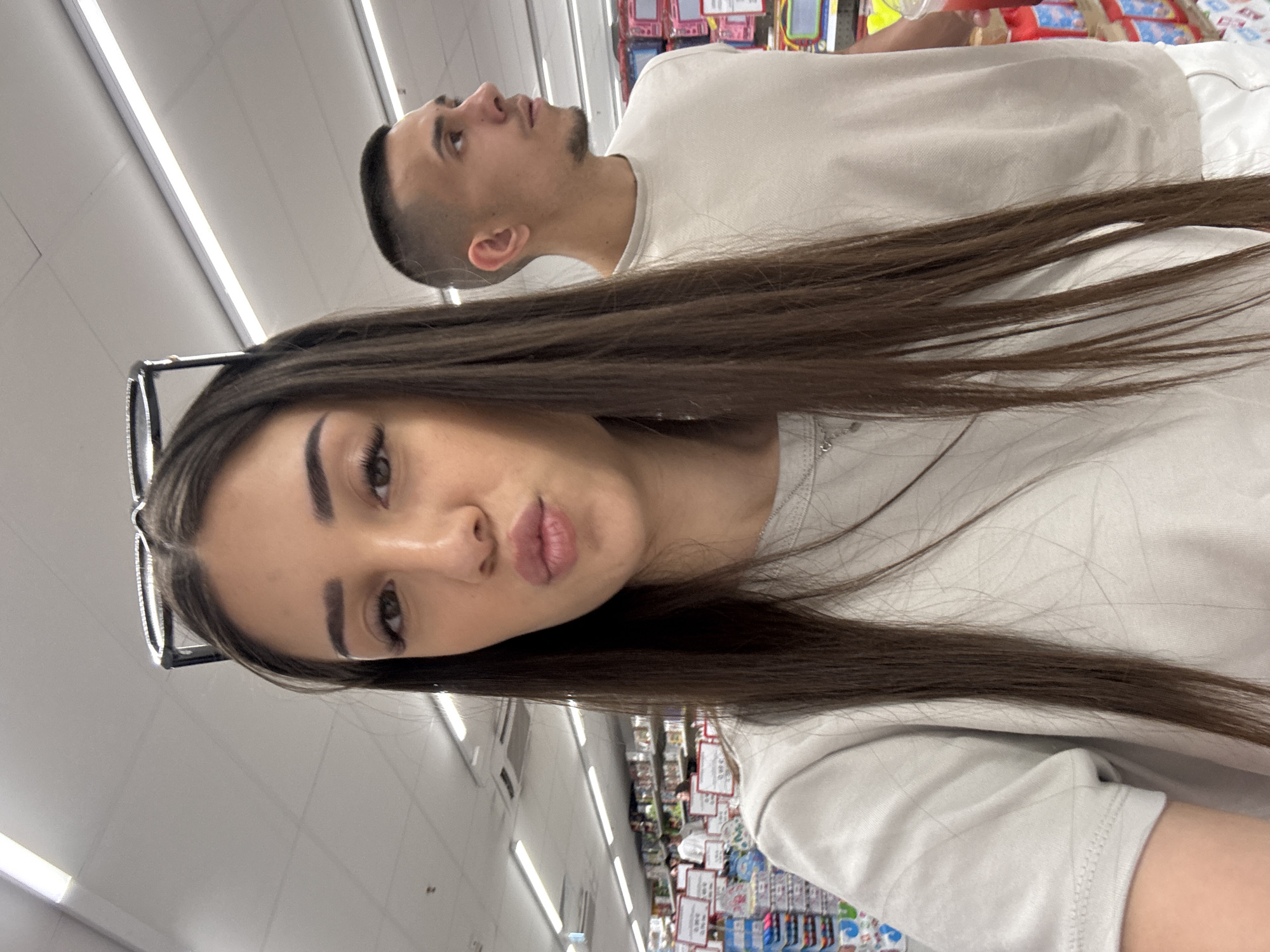 This image captures a selfie taken inside a retail store located in Plovdiv, Bulgaria. In the foreground, a young woman with long, straight, dark brown hair, parted centrally, poses for the camera. She wears sunglasses perched on top of her head and a light grey short-sleeved t-shirt with a silver chain necklace visible. Her lips are pursed in a "duck face" expression, and her dark eyebrows and prominent eyelashes are visible. Behind her and to the right, a young man with a short fade haircut and light stubble looks to his right (viewer's left), appearing disengaged from the camera. He wears a light beige or off-white t-shirt. The background reveals rows of retail shelving stocked with various products, some with white signs featuring red text, suggesting prices or product information. The store's ceiling is light-colored and features multiple rectangular fluorescent light fixtures and an air conditioning vent. A red cup is partially visible on the far right, possibly held by the man.