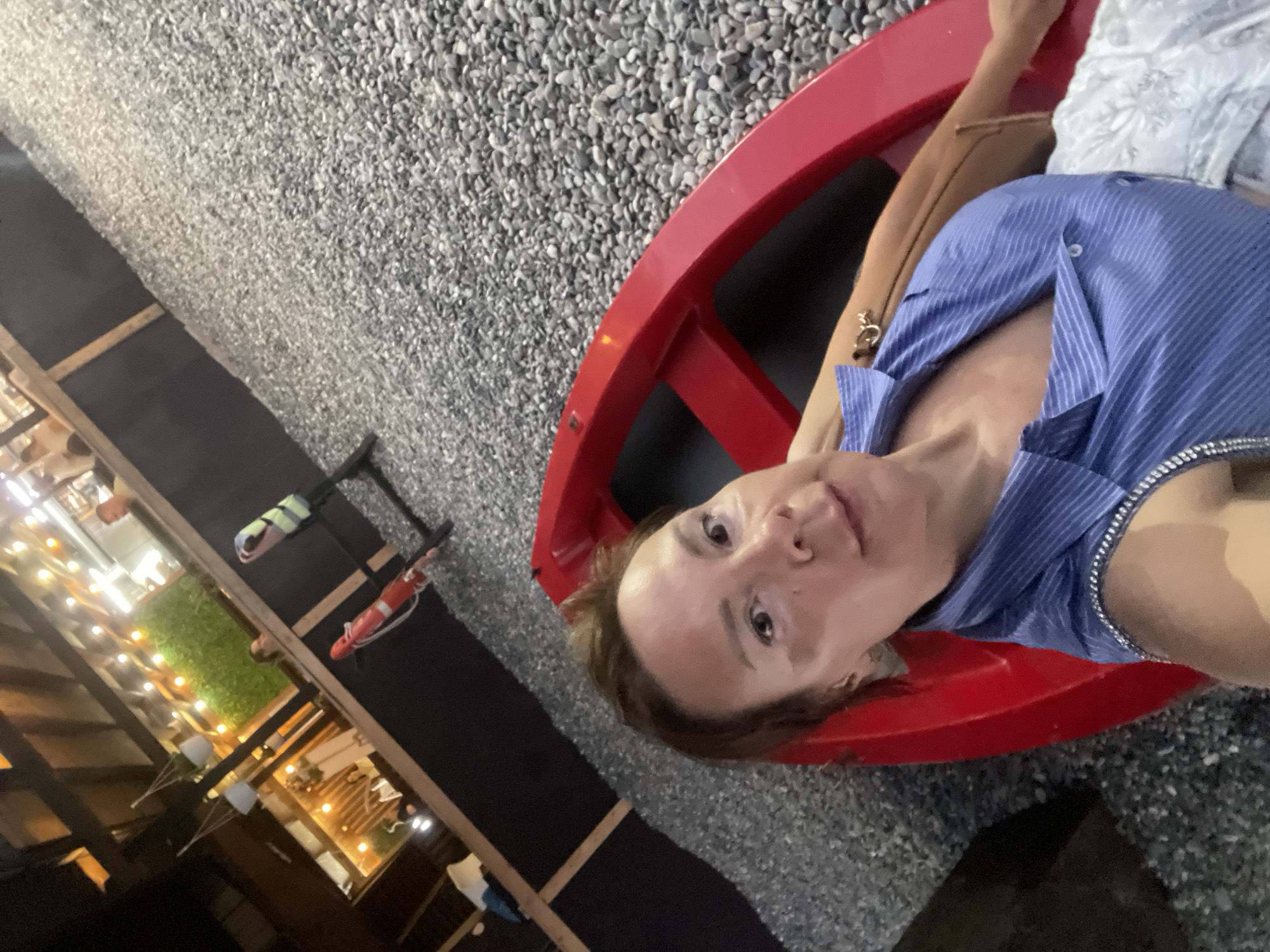 A woman is lying in a bright red boat on a gravelly surface. She is wearing a blue striped shirt and has brown hair. The boat is positioned at an angle, with the woman's face closest to the camera. In the background, there is a structure with lights strung across it, and some greenery. There are also some safety buoys visible. The scene appears to be outdoors, possibly in a recreational area or a restaurant. The lighting suggests it is evening or night.