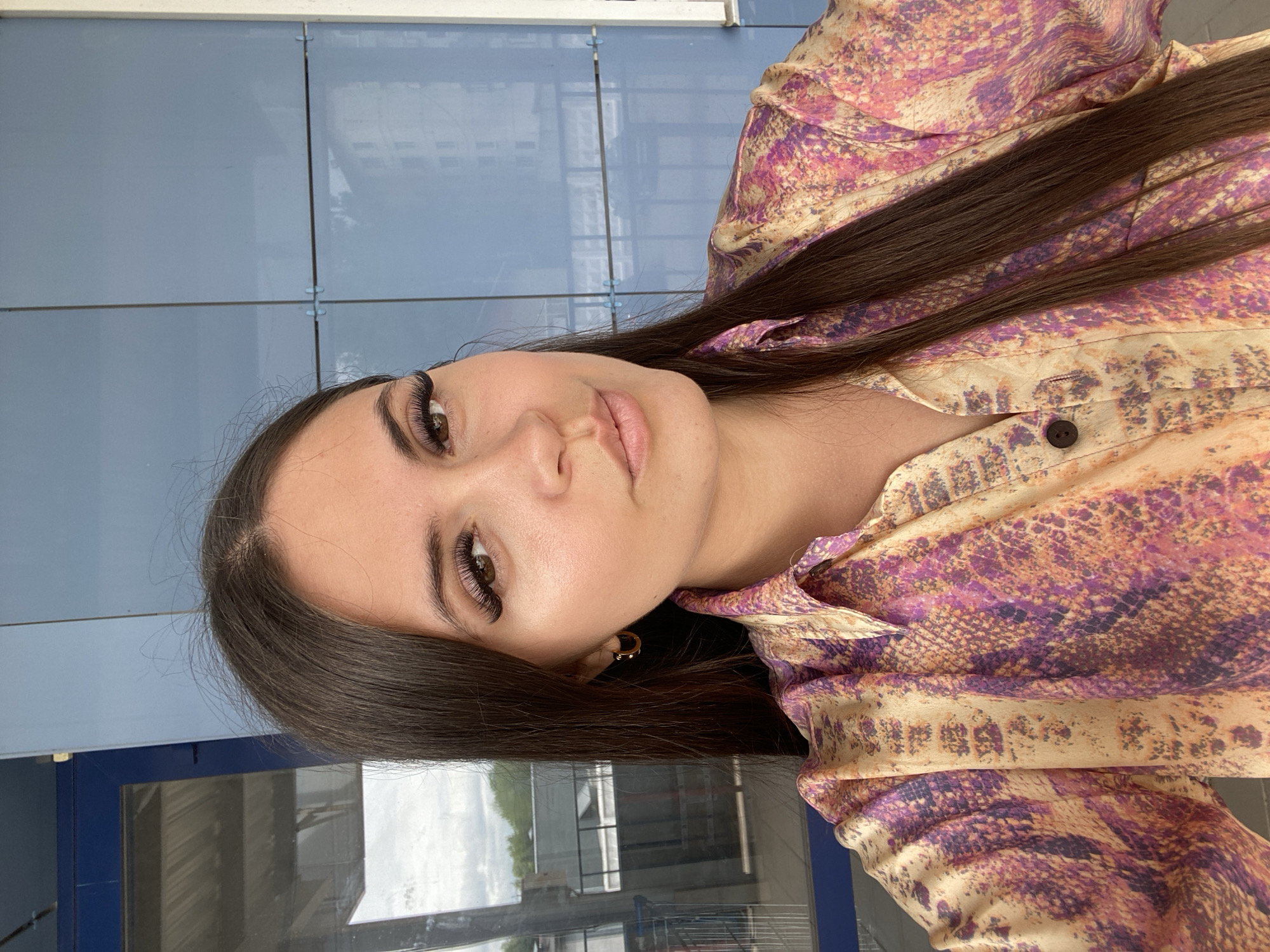 A close-up, eye-level, outdoor, waist-up selfie shows a young woman with long, dark brown hair, wearing a colorful button-up shirt with a snake-like pattern. She has on large, dramatic false eyelashes and small gold hoop earrings. The background consists of a light blue wall with glass panels, reflecting buildings and trees. The woman appears to be standing on a platform or walkway, possibly at a train station or public transport hub, given the presence of a metal railing visible in the background reflection. The lighting is natural and appears to be daytime. The image was taken on the outskirts of Chaikovsky, Russia.