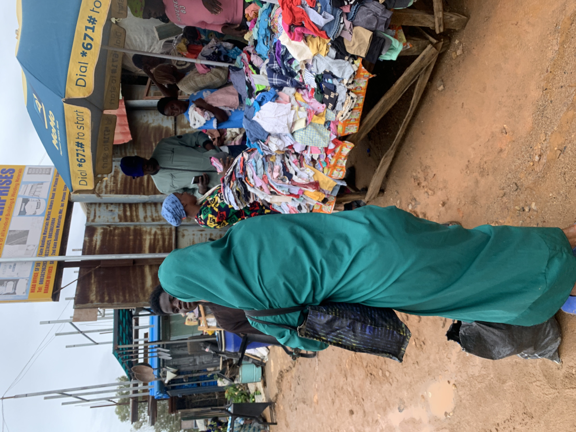 A street scene in Gura Top, Nigeria, presents a vendor's stall piled high with an assortment of colorful clothing. A blue umbrella bearing the mobile money company "MOMO" logo provides shade to the vendor and customers. Several individuals are present; some appear to be examining the clothes, while one person in a green garment stands in the foreground. The ground is dirt and mud, suggesting a recent rain or unpaved conditions. A sign for "Enterprise", listing telephone numbers and branch offices with a construction material focus is on the left. Steel and building materials can be seen in the background behind the vendor's stall.