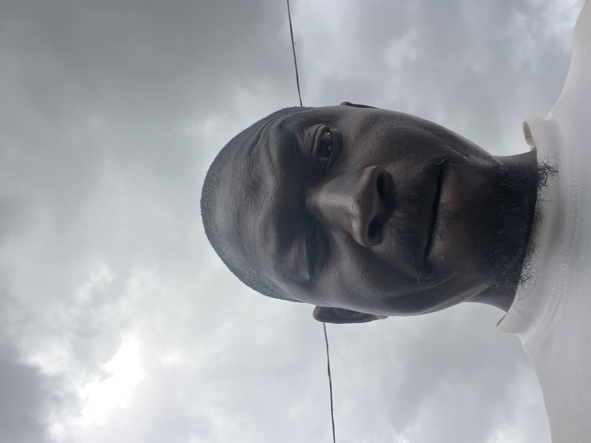 The image is a close-up, low-angle, outdoor selfie of a dark-skinned man. He is looking directly at the camera, with his head tilted slightly. His left eye is partially closed, and his right eye is open. He has a goatee and a mustache. He is wearing a white, high-necked shirt. A thin, dark wire runs horizontally across the frame, behind the man's head. The background is a cloudy, overcast sky. The image appears to have been taken in Jos, Nigeria, on a day with heavy cloud cover.