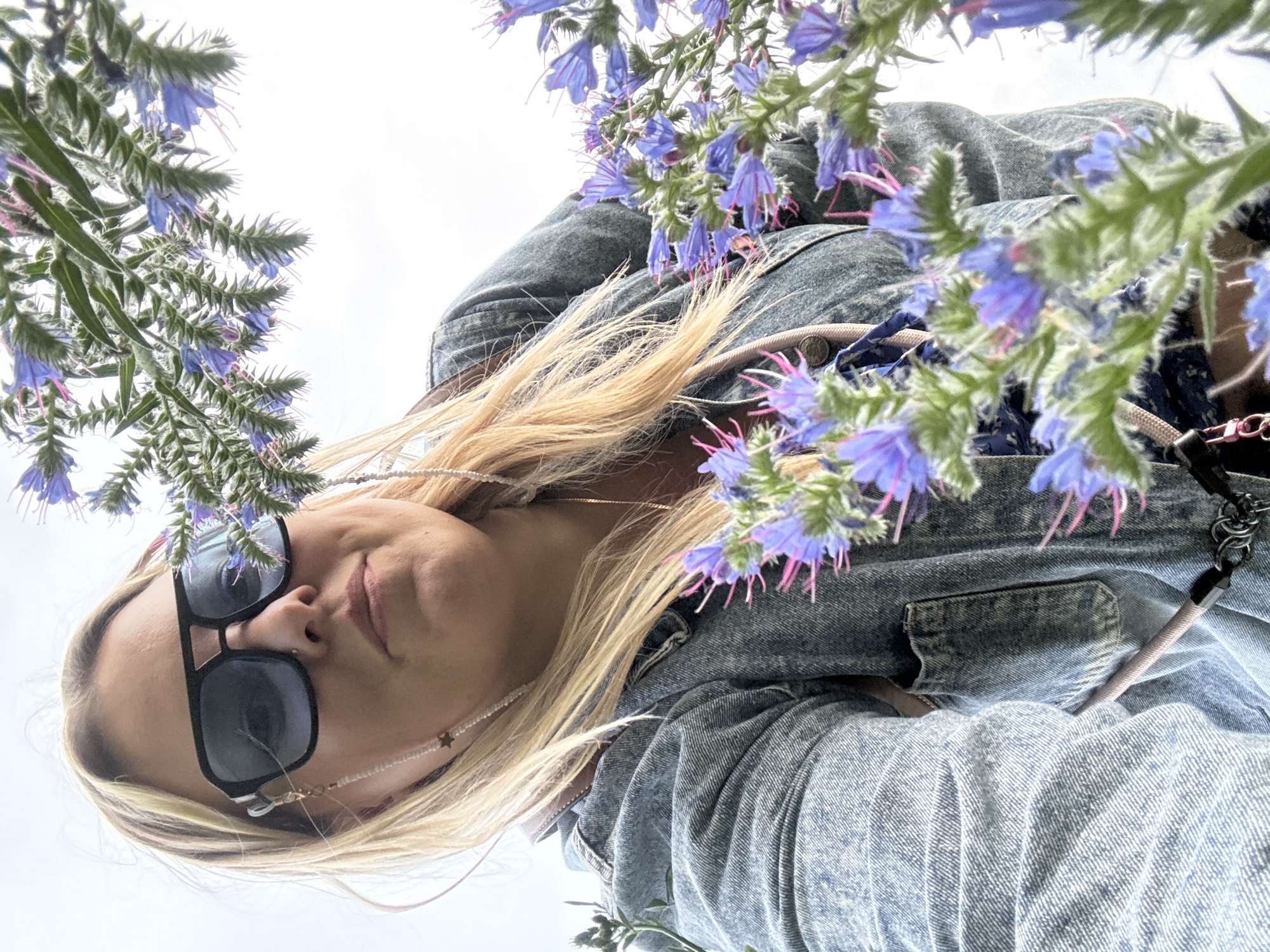 This is a low-angle, portrait-oriented, upward-facing photograph of a young woman. She has blonde hair and is wearing sunglasses and a denim jacket. The woman is framed by blue flowers with pink accents, which appear to be growing on tall, fuzzy stems. The sky, a bright white, suggests it is daytime, likely under overcast or bright conditions. The woman's head is tilted back, and she has a slight smile, conveying a sense of contentment or enjoyment. The background is mostly the bright sky, with some of her blue denim jacket visible, including a pocket on the right side. The image seems to have been taken outdoors, possibly in a field or garden, given the presence of the flowers. The orientation of the photo suggests the photographer was lying down or at a very low vantage point. There is no visible text in the image. The context provided places the scene in Марковский, Russia, though no specific landmarks confirm this. The overall mood is relaxed and cheerful, with a focus on the woman and the surrounding nature.