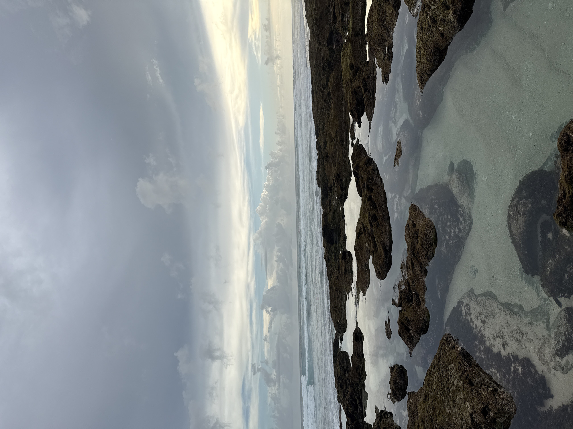 The image depicts a coastal scene in Pecatu, Indonesia. The foreground consists of dark, jagged rocks interspersed with shallow pools of water, the clear water reflecting the sky above. Beyond the rocks, the ocean meets the horizon, showing some waves. The sky is overcast, with a mix of dark gray and white clouds. The scene appears to be captured during daylight, possibly at dawn or dusk, given the soft lighting. There are no people or human-made structures visible in the frame.