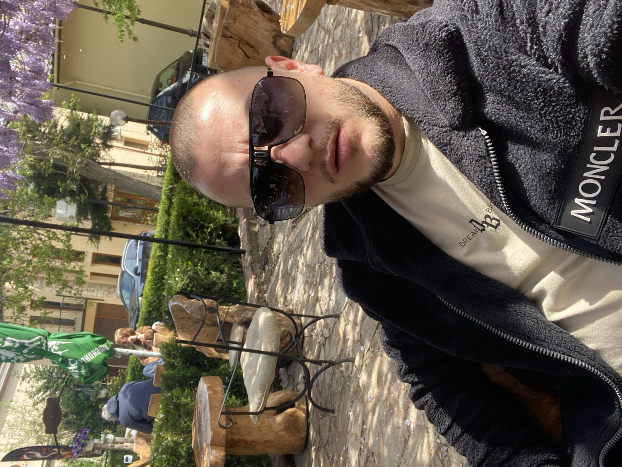 The image shows a man in a black fleece jacket with a "MONCLER" label, wearing sunglasses and a beige t-shirt with the word "DREAMER" on it. He is sitting on a wooden chair in an outdoor setting. In the background, there are other people seated, green umbrellas, trees, and a building. Based on the vegetation and the style of the outdoor furniture, it is likely the image was taken in Varna, Bulgaria.