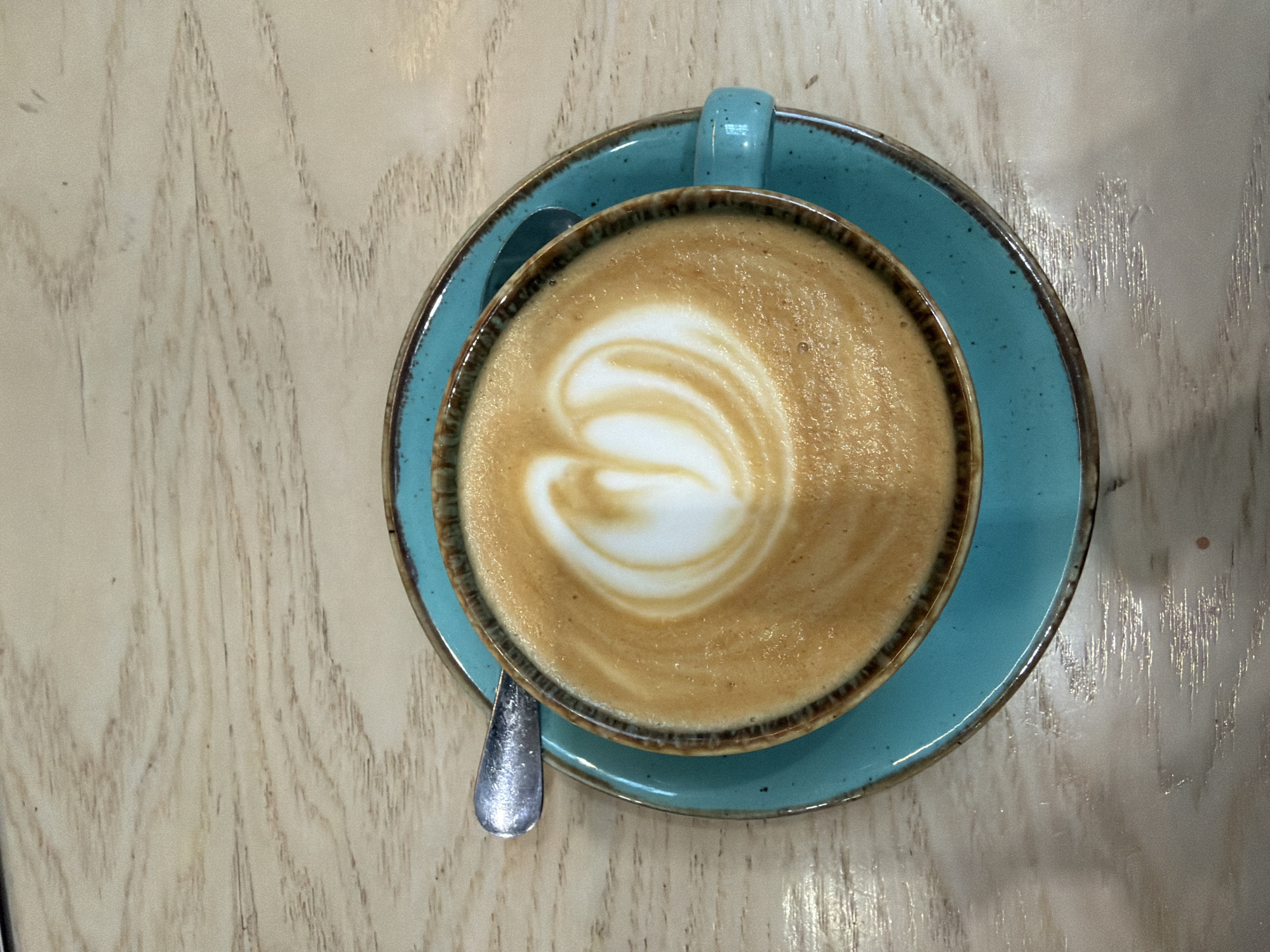 The image shows a top-down view of a cup of coffee with latte art on a blue saucer. A silver spoon rests on the saucer, beside the cup. The setup is placed on a light-colored wooden table. The location is not specified.