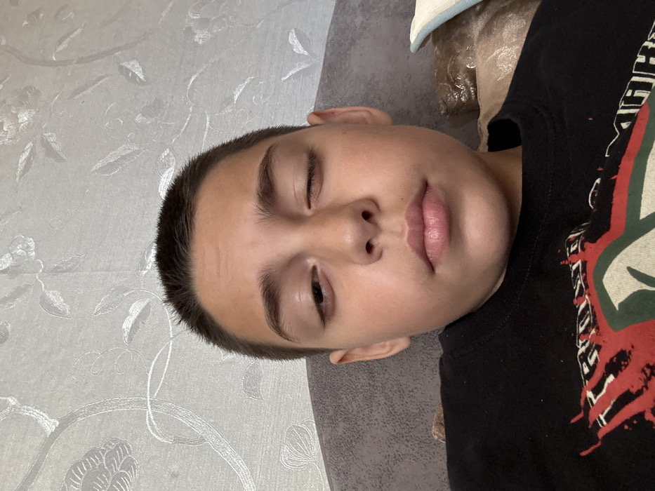 An indoor close-up depicts a young individual with short, dark hair and a light complexion, situated in what is reported as the city of Chervena voda, Bulgaria. The subject faces the camera directly. The left eye (viewer's left) is partially open, while the right eye (viewer's right) is closed. The mouth is closed, maintaining a neutral facial expression. The individual wears a black crew-neck t-shirt displaying a partial graphic design in red and white on the chest. The background features a light-toned wall covered in wallpaper with a subtle, embossed floral or vine pattern. Portions of a dark grey cushion or headrest are visible on the left, and a beige, possibly plastic-covered cushion along with a light-colored striped cushion are visible on the right.