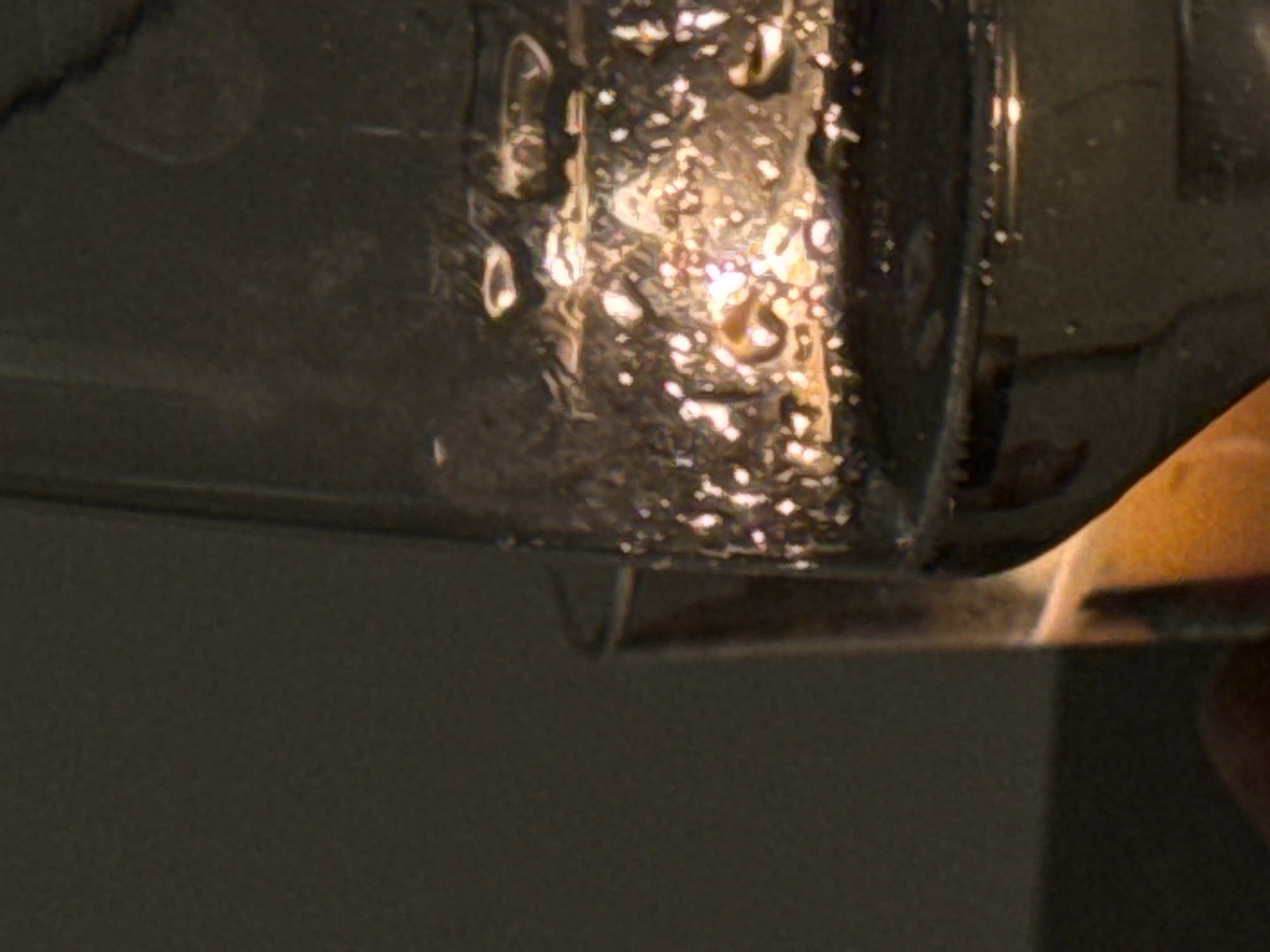 The image shows a close-up of a dark, likely glass bottle with water droplets condensing on its surface. The drops reflect light, creating bright highlights. The bottle appears to be positioned near a metallic surface. A section of what appears to be a human hand is visible. The background is plain and dark.
There is no information to determine the location in the city of Munich, Germany.