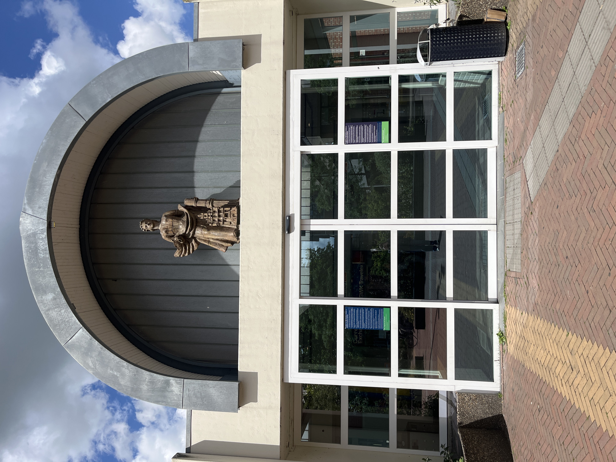 A front-facing, eye-level outdoor shot shows the entrance to a building with automatic glass doors. Above the doors, a large, dark gray, arched facade is visible. Centered within the arch is a bronze sculpture of a figure holding a smaller building. The building facade is painted a light cream color. The automatic doors are made of white-framed glass panels. A dark gray diamond-patterned trash can is positioned to the right of the entrance. The ground in front of the entrance is paved with reddish-brown bricks laid in a herringbone pattern, with a yellow brick stripe running diagonally across it. The sky above is blue with scattered white clouds. The scene appears to be in a city, but the specific location is unknown.