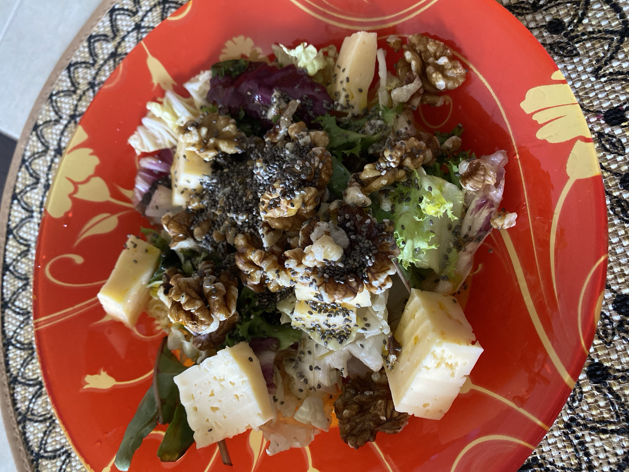 A close-up, overhead shot shows a salad in a red, decorative bowl. The salad is composed of various types of lettuce, chunks of white cheese, walnuts, and chia seeds. The bowl itself has a glossy finish and features a yellow, floral-like pattern. The bowl rests on a textured placemat with a black and white pattern. The scene is well-lit, likely from natural light, and there is no indication of the specific location within Lida, Belarus.