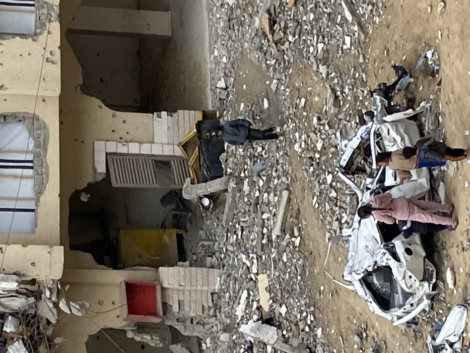 The image depicts a scene of extensive destruction in what appears to be a residential area in Khan Yunis, Palestinian Territory. The ground is covered with significant amounts of rubble, broken concrete, and other debris.
A multi-story building on the left side of the frame is severely damaged, with large sections of its walls missing, exposing interior spaces. Numerous small, circular holes are visible on its exterior walls, suggesting bullet or shrapnel impacts. Windows are shattered or completely absent. Parts of the building's facade appear to have crumbled.
In the center-right of the frame, a white car is seen, completely mangled and crumpled, indicating severe damage, possibly from an explosion or collapse. Its roof and sides are caved in.
Several people are present amidst the devastation. One individual, dressed in a dark jacket and pants, stands in the middle distance near the damaged building, seemingly surveying the area. Closer to the wrecked car, three other individuals are visible. One is wearing a pink outfit, another a brown or tan top, and a third a blue top, appearing to be children or young adults, standing near the severely damaged vehicle.
Other objects include a partially visible yellow container or structure near the damaged building, and a red container or object closer to the foreground. The scene is illuminated by daylight, with no strong shadows, suggesting an overcast day or midday light. No discernible text is visible in the image. The overall impression is one of widespread devastation and aftermath.