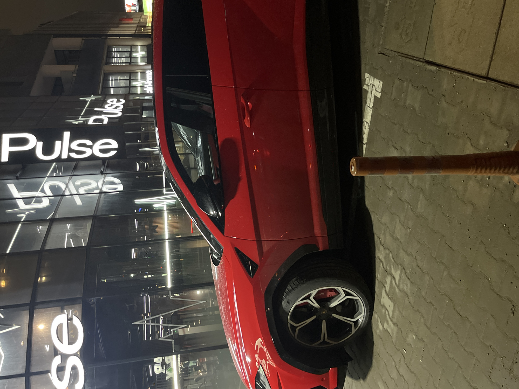 A red Lamborghini Urus is parked in front of a building with a large "Pulse" sign in Sofia, Bulgaria. The vehicle is positioned on a paved surface, next to a metal bollard. The building behind the car features a glass facade with the "Pulse" signage prominently displayed. The environment appears to be urban, likely a commercial area. No individuals are visible in the image. The image is taken at night.