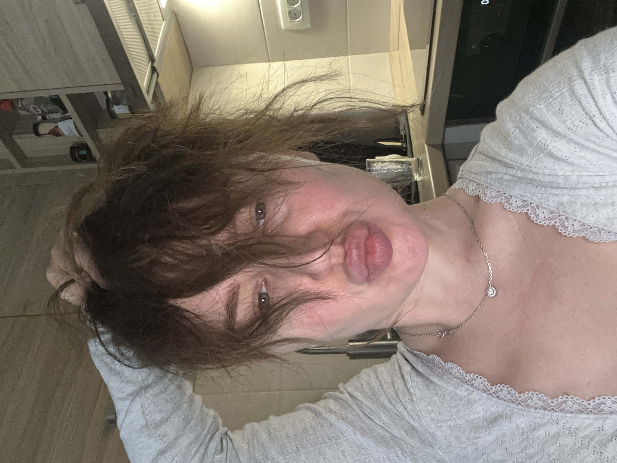A female subject is depicted in a close-up, head-and-shoulders selfie taken in a modern kitchen setting within Budva, Montenegro. Her unkempt, light-brown hair, with strands falling across her face, is partially held by her right hand, which is raised above her head. Her face exhibits some redness, and her lips are pursed in a "kissing" expression. Her visible left eye is directed towards the camera. She wears a light-grey, textured knit top featuring a white lace trim along the neckline and a silver-colored necklace with a small, circular, faceted pendant. The background reveals light wood-grain patterned kitchen cabinets, one shelf holding various bottles and jars. Below the cabinetry is a stainless steel range hood. To the right, a light-tiled backsplash with a white double power outlet is visible. A dark countertop holds a clear, textured glass. A dark appliance, likely an oven or microwave, with a partially visible digital display, is seen below the countertop. The scene is illuminated by artificial, warm lighting.