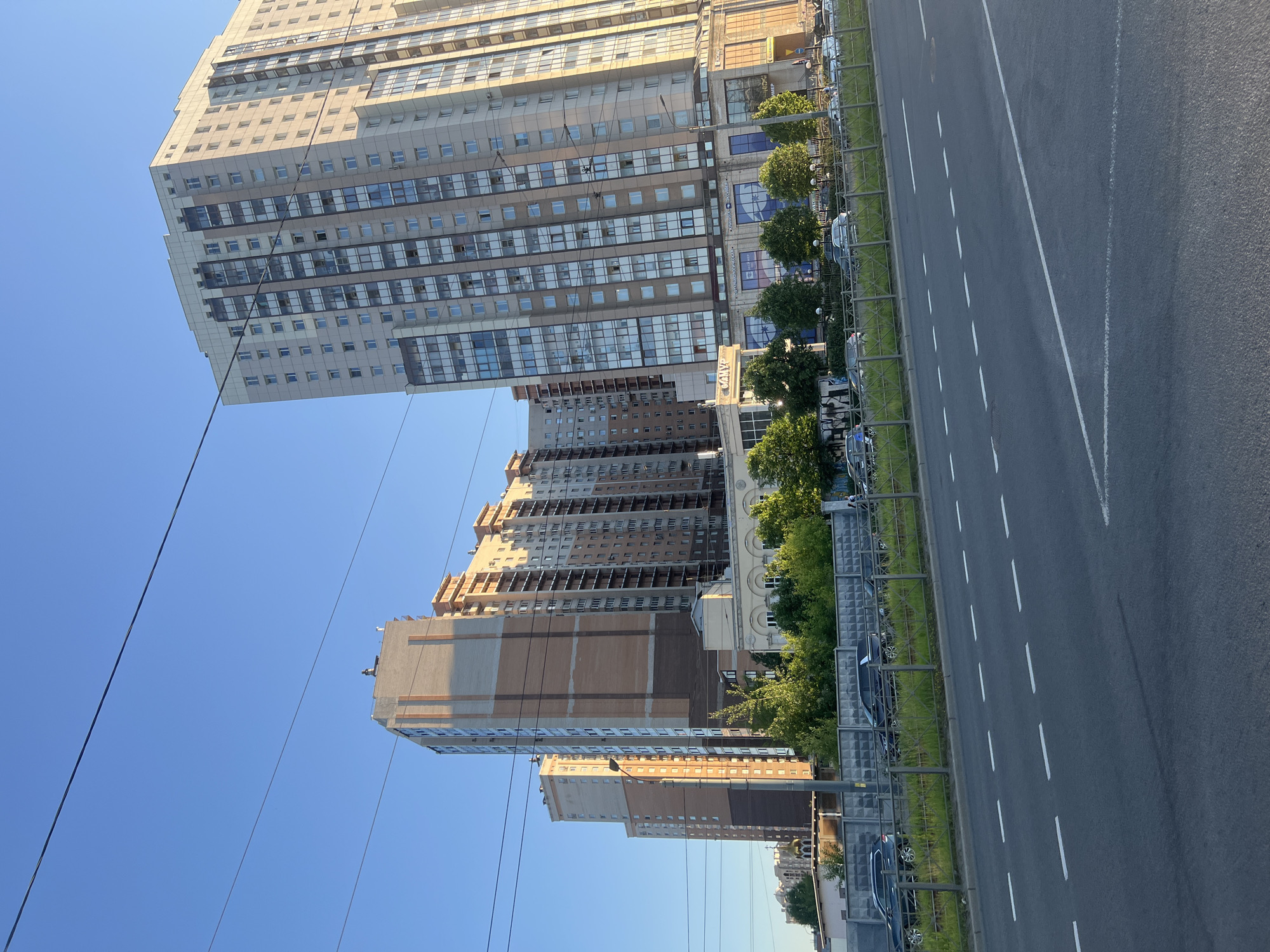 This detailed description pertains to an urban street scene captured during daytime in Saint Petersburg, Russia, specifically on Ulitsa Bely Kuna, looking towards the multi-story residential and commercial complex at Ulitsa Bely Kuna 3.
The foreground features a wide, dark asphalt road with two parallel sets of dashed white lane markings, indicating multiple lanes of traffic. The road surface appears dry and clear.
Immediately beyond the road, a low metal fence with a diamond-patterned mesh runs horizontally across the mid-ground. Behind this fence, a strip of green grass and densely packed green trees and bushes are visible. Further back, a long, light gray concrete wall with a rectangular block pattern extends across the scene. Several dark-colored passenger vehicles, including SUVs and sedans, are parked parallel to this wall and fence. On the far left, partially obscured by the wall and trees, a golden onion dome, characteristic of an Orthodox church, is visible.
The background is dominated by several large, multi-story residential buildings under a clear, bright blue sky.
On the right, a prominent, tall residential building features a facade of light beige and cream panels, interspersed with vertical sections of darker brown and gray cladding. It has numerous rectangular windows and some balconies. The ground floor houses commercial establishments with large glass storefronts displaying various signs in Cyrillic, including "КЛИМАТ" (Climate), "ПРИМЕР" (Example/Sample), and a blue circular logo. This building is bathed in warm, golden sunlight, suggesting either early morning or late afternoon.
To the left of this building, slightly set back, stands another tall residential structure with a lighter brown and beige facade. Its lower section incorporates classical architectural elements, including arched windows and columns, above which a sign reading "САМ" (likely part of "САМПО") is visible.
Further to the left and receding into the background, additional multi-story residential buildings with similar architectural styles are present.
Several thin, dark electrical or communication wires traverse the blue sky horizontally across the upper portion of the image. A few indistinct human figures are discernible on the sidewalks or near the commercial establishments, but their actions or specific features are not clear. The overall environment is one of a modern, developed urban area.