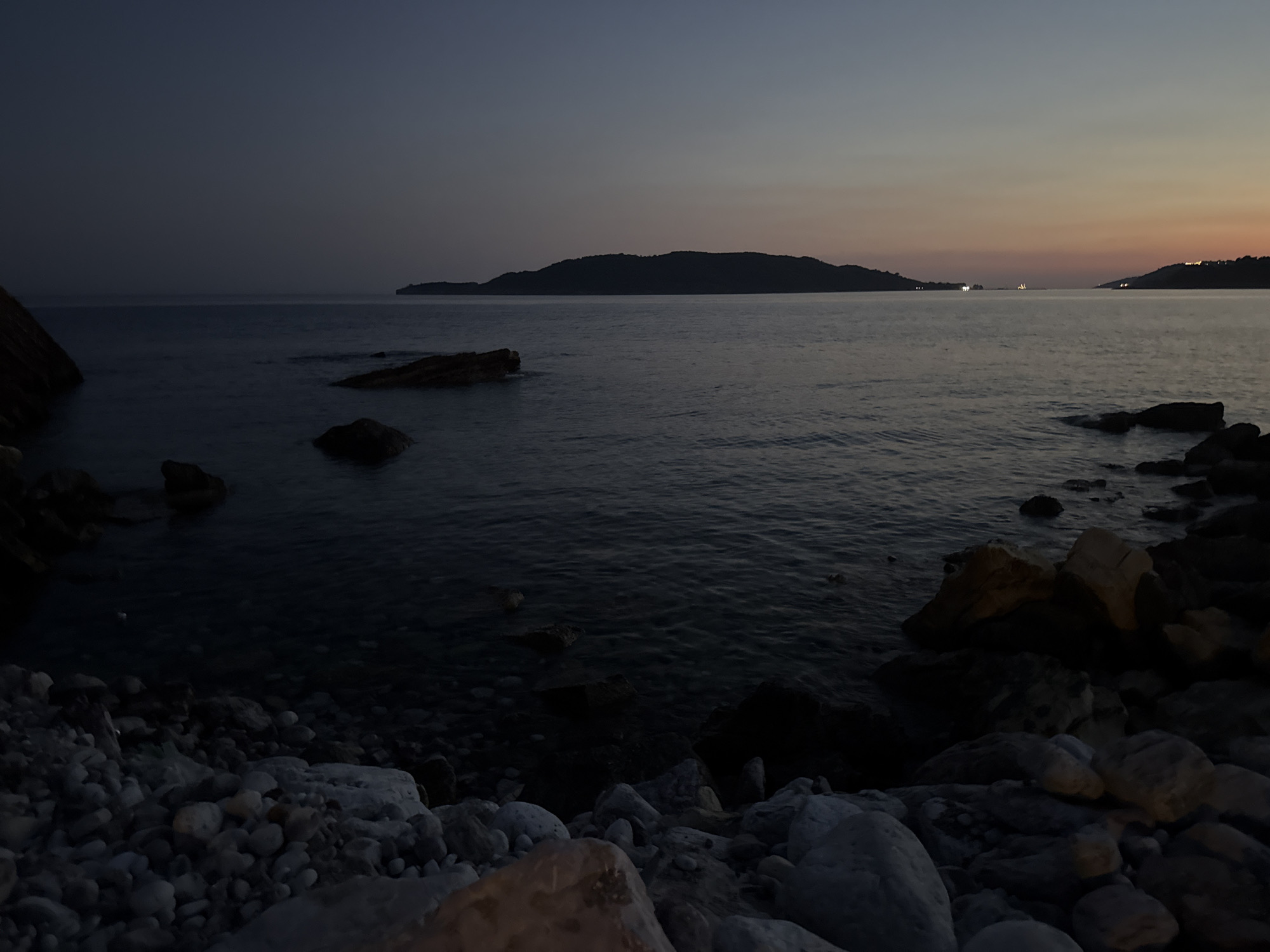 The image depicts a twilight scene at a rocky shoreline in Bečići, Montenegro. The sky is a gradient of dark blue transitioning to pale orange and pink hues near the horizon, indicating the sun has recently set. The sea is calm with gentle ripples, reflecting the muted colors of the sky.
In the distance, a silhouetted island or peninsula stretches across the horizon, its form indistinct against the fading light. A few small lights, possibly from boats or distant buildings, are visible on the horizon line.
The foreground is dominated by a rocky beach composed of various sizes of stones and pebbles, with larger rocks interspersed along the water's edge. The water is dark and deep, with some rocks visible beneath its surface. The overall atmosphere is serene and tranquil, characteristic of a coastal landscape at dusk. There are no people or discernable activity in the scene.