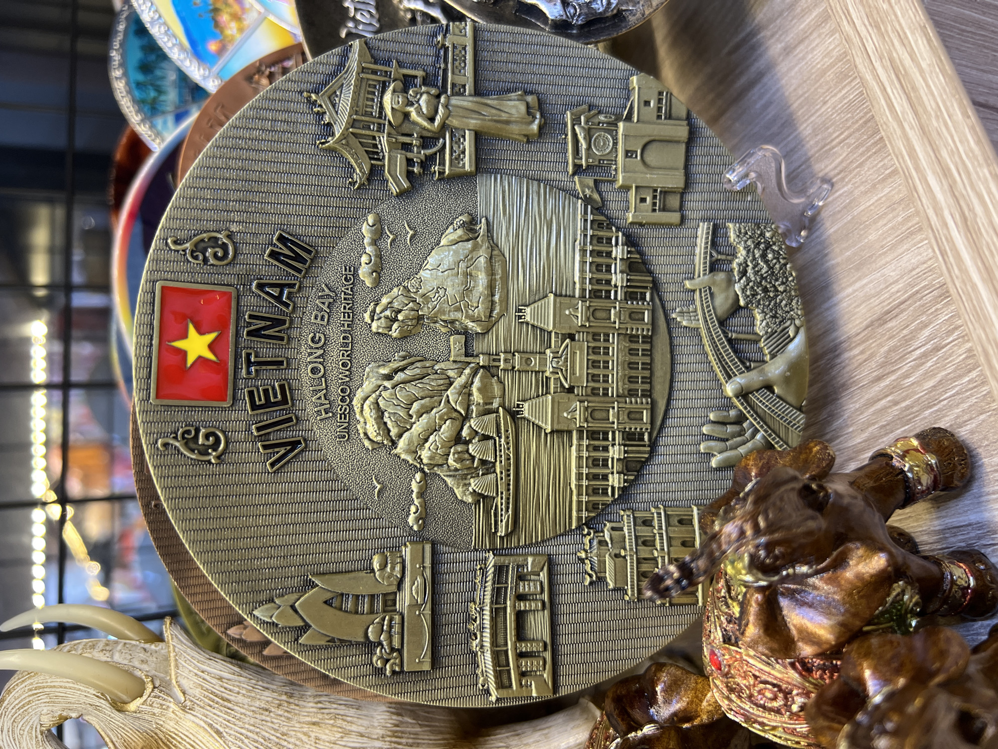 The image shows a close-up of a souvenir plate or disk depicting landmarks and scenery from Vietnam. The plate is primarily gold-colored and features a raised relief design. Prominently displayed at the top is the Vietnamese flag with a yellow star on a red background. Below the flag, the word "VIETNAM" is written in bold, raised letters. Further down, "HALONG BAY" and "UNESCO WORLD HERITAGE" are inscribed. The central motif of the plate depicts iconic limestone karsts rising from the water, with traditional Vietnamese dragon boats sailing between them. Scattered around this central image are smaller reliefs of various architectural structures, including pagodas, temples, and grand buildings. A traditional Vietnamese woman in conical hat and long dress is depicted on the right side of the plate. In the foreground, partially obscuring the bottom portion of the plate, are several decorative elephant figurines. The elephants are brown and appear to be adorned with colorful, ornate coverings. The plate is resting on a light-colored wooden surface, and the background shows a blurred view of other souvenirs and a display shelf with lights. The scene appears to be within a retail environment, likely a souvenir shop. There is no indication that this specific location is in Nha Trang, Vietnam; the plate depicts general Vietnamese heritage sites.
