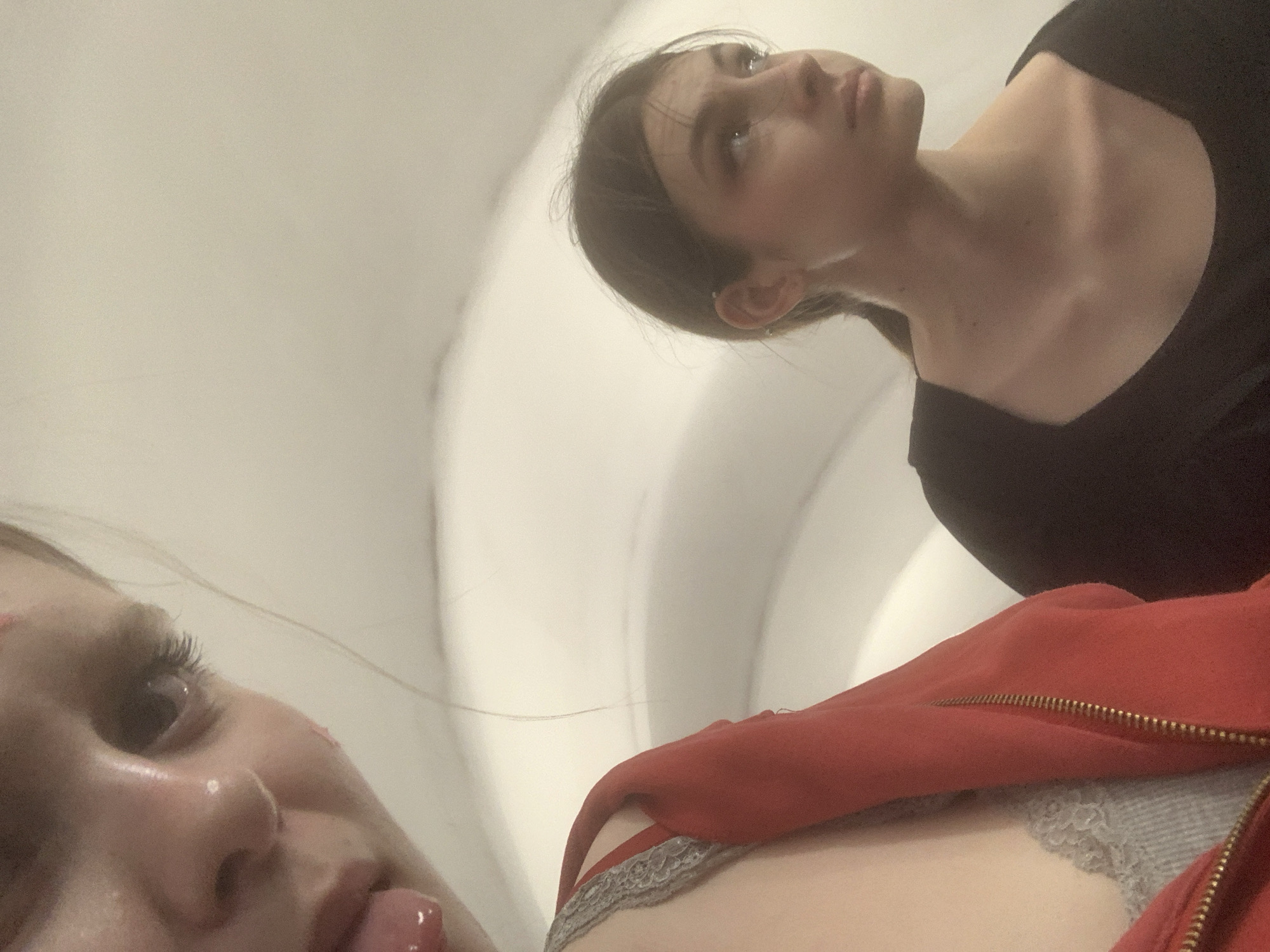 The media file is a low-angle, candid portrait featuring two young women in what appears to be an indoor setting in Saint Petersburg, Russia.
In the upper right portion of the frame, one woman is visible from the chest up. She has dark hair, pulled back, and is wearing a dark, possibly black, top. She looks upwards and slightly to the right with a calm, somewhat pensive or observant expression. Her neck and collarbone are clearly defined.
In the lower-left foreground, partially out of frame and very close to the camera, is the face and shoulder of a second woman. Her eye is open and also looking upwards, with a glisten visible near her nose, which could suggest moisture or a tear. Her mouth is slightly open, revealing part of her tongue. She has lighter hair, with some strands visible across her face. She is wearing a red zippered garment, possibly a hoodie or jacket, over a grey, lace-trimmed top or bra.
The background is a soft, off-white, minimalist interior space with subtly curved surfaces, illuminated by diffuse ambient light. The low camera angle and close-up of the foreground subject create an intimate and immediate perspective. Both individuals appear to be looking in the same general direction, suggesting a shared point of focus or observation. There is no visible text in the image.