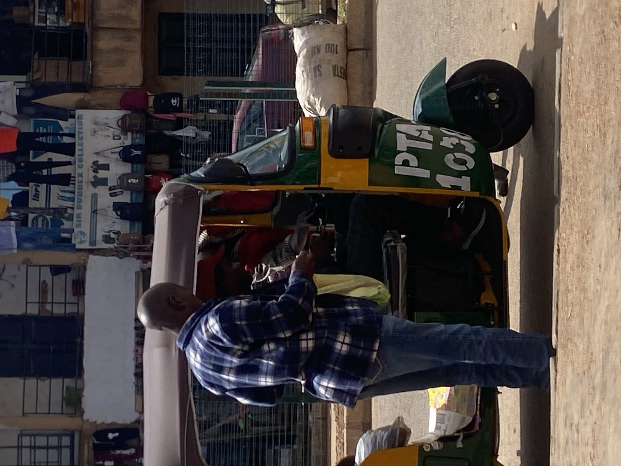 This image captures a daytime street scene in Bukuru, Nigeria. In the foreground, a bald, dark-skinned male, dressed in a blue and white plaid long-sleeved shirt and blue jeans, stands next to a three-wheeled auto-rickshaw. He is positioned on the left side of the vehicle, facing towards its interior, holding a light-colored plastic bag in his left hand, and interacting with a partially visible individual inside the vehicle whose right hand is extended. The auto-rickshaw is primarily green and yellow, with a dark canopy, and has "PTA 1030" inscribed in white text on its green side panel.
In the mid-ground, to the right of the auto-rickshaw, a large white sack with "ETA SA 100 KU" printed on it rests on a low concrete barrier or curb. A dark red car is partially visible further down the street behind the auto-rickshaw. The background features several buildings. A building on the left exhibits dark window grates and a white painted section below. Centrally, a commercial establishment is visible with a sign displaying "SIR DUBBEX COLLECTION" and "lhri" above. Various items of clothing, including shirts and jeans, are displayed, along with multiple backpacks and bags of different colors, hanging below the main sign. The ground is a light-colored paved surface, and distinct shadows indicate bright sunlight.