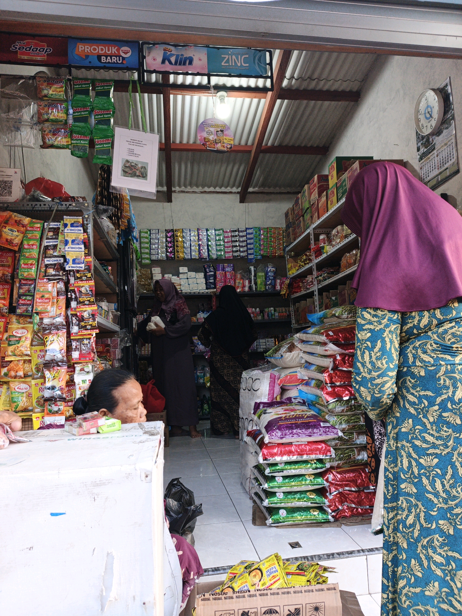 The image depicts the interior of a small convenience store in Ketegan, Indonesia. The store is packed with merchandise, including rows of packaged food items, snacks, and cleaning supplies displayed on shelves. A woman wearing a purple hijab and a patterned blue and gold dress stands in the foreground, facing away from the camera, next to stacks of rice bags. To her left, another woman wearing a dark hijab is visible among the shelves, holding a white item. In the lower left corner, a woman with dark hair pulled back is leaning over a white counter, looking to the right. The store's corrugated metal roof is visible, along with hanging signs advertising various brands. A clock on the wall shows approximately 10:20. The overall impression is of a busy, well-stocked neighborhood store.