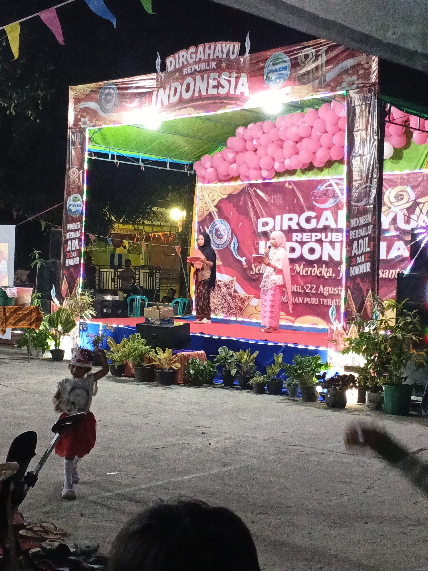 A stage is set up for an event in Cikande, Indonesia, likely a celebration of Indonesia's Independence Day, given the banner that reads "DIRGAHAYU REPUBLIK INDONESIA" (Happy Independence Day Republic of Indonesia). The stage is decorated with pink balloons and colorful lights. Two women are on the stage, one on the left holding a microphone and a piece of paper, and another on the right holding a paper. A young girl wearing a red skirt and a white top with a hat is in the foreground, looking towards the stage. Various potted plants are arranged around the stage. The event appears to be taking place outdoors in an open area, possibly a public square or community gathering space. The lighting suggests it is either dusk or nighttime.