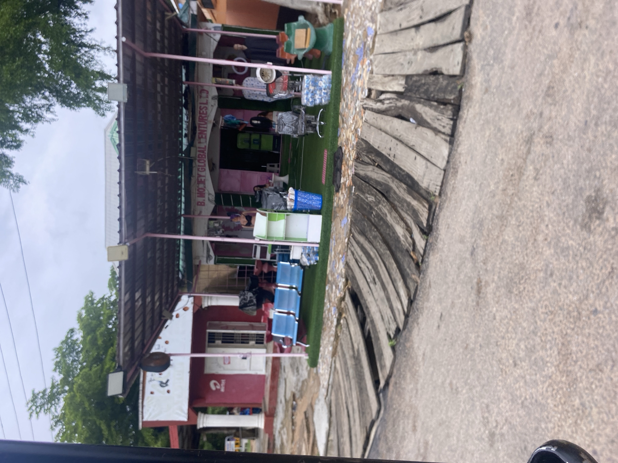The image captures a street-level view in Jalingo, Nigeria, likely on a cloudy day given the overcast sky and soft lighting. In the foreground, a rough, weathered wooden walkway with uneven planks spans across the frame, leading towards a commercial establishment. The walkway appears damaged and aged, with some planks askew.
The commercial building features a prominent pink facade with white pillars. Above the entrance, a sign reads "B. MOJEY GLOBAL VENTURES LTD." The building is covered by a dark, corrugated metal awning supported by pink posts. Artificial turf covers the ground in front of the shop, creating a designated outdoor display area.
Several objects are arranged on the turf, including blue plastic chairs, a green shelving unit with some items, a black office chair, stacks of bottled water, and a green, cartoon-like sculpture resembling a character. Mannequins are also visible, some dressed in clothing. The shop interior appears to be open, with glimpses of what could be more merchandise or seating.
To the left of the main shop, another section of the building, painted red, displays an Airtel logo. A person can be seen partially through a glass door in this section. The overall scene suggests a busy commercial area, possibly a market or a street lined with shops, with evidence of wear and tear in the surrounding infrastructure. The presence of both a wooden walkway and a paved surface indicates a mix of urban development and potentially less maintained areas.