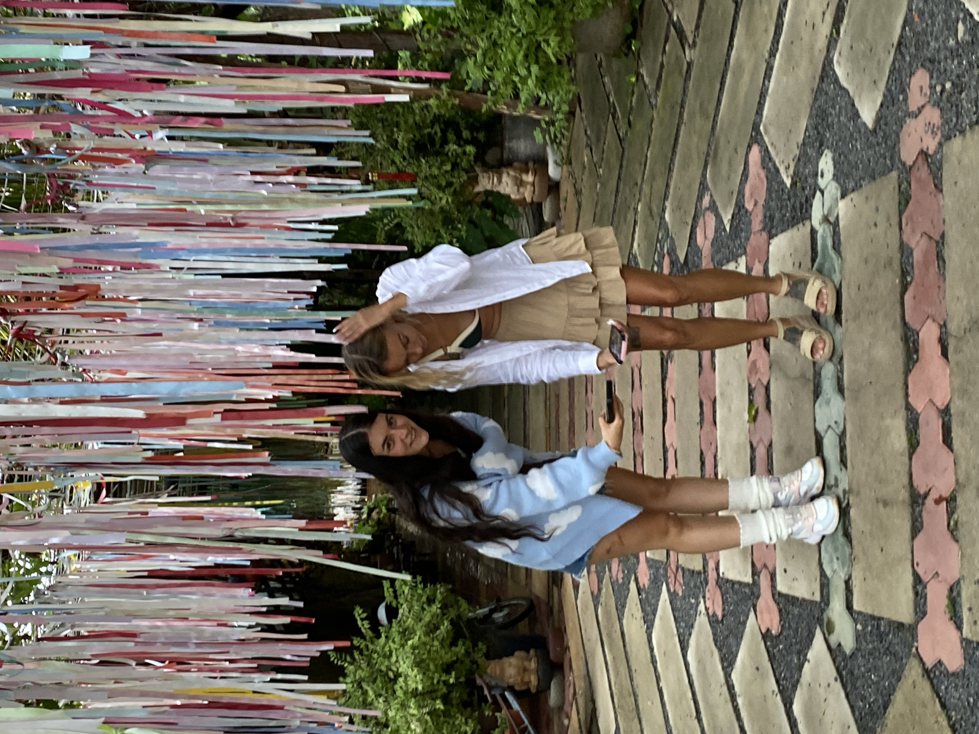 Two young women are standing side-by-side outdoors on a paved walkway. The woman on the left has long, dark hair and wears a blue sweater with white cloud patterns and white socks with sneakers. She is holding a phone, seemingly taking a selfie. The woman on the right has blonde hair and wears a white button-down shirt over a tan dress and sandals. Multi-colored ribbons hang overhead, creating a canopy. Green vegetation surrounds the paved area. The setting is Mũi Né, Vietnam, based on the distinct paved walkway design.