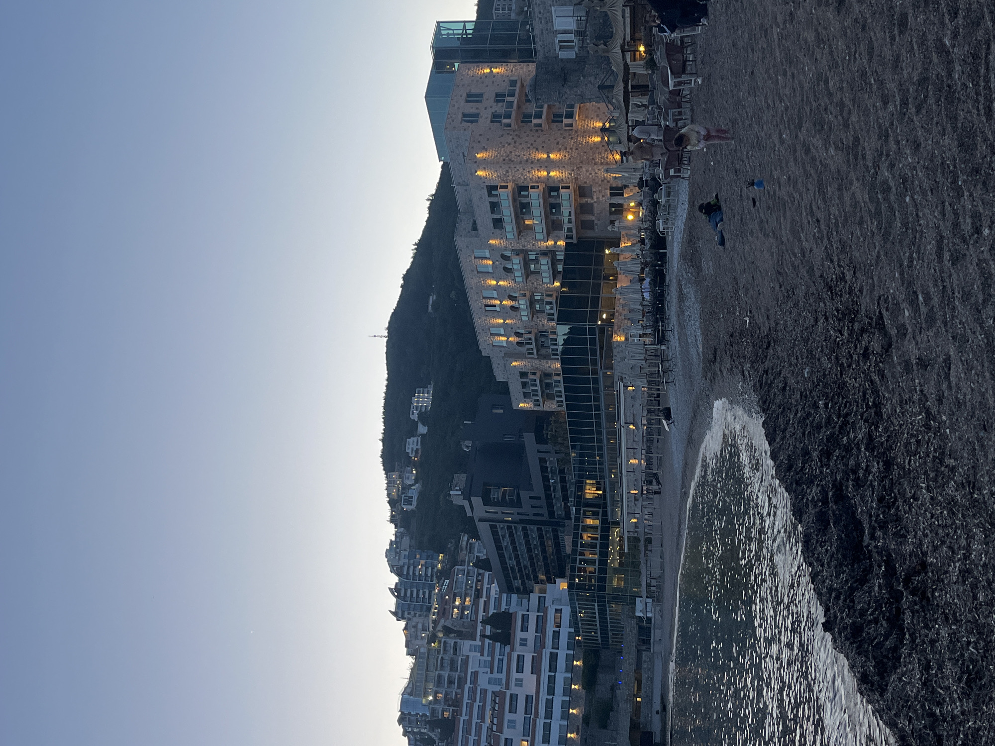 The image shows a coastal cityscape at dusk, with a beach in the foreground and a cluster of buildings rising up a hillside. The city is unknown.
The beach is dark and coarse, with waves lapping at the shore. People are present on the beach, including an adult standing near beach chairs and a person lying down.
Buildings vary in height and architectural style, with some newer, modern structures and others appearing older and more traditional. Lights are visible in the buildings, suggesting it is near sunset.
The sky is a pale blue, indicating the time of day. A hillside rises behind the buildings.