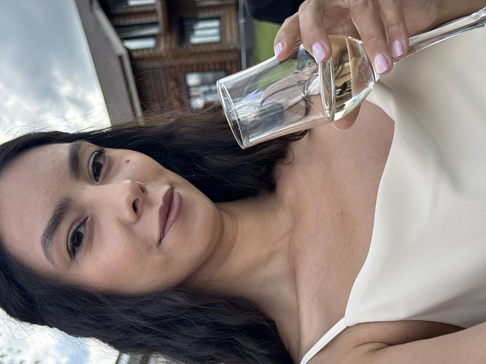 A young woman with dark, wavy hair is holding a glass of champagne and looking at the camera. She is wearing a light-colored dress. In the background, there is a large wooden house and trees. The sky is cloudy. The image appears to have been taken outdoors in a rural setting, potentially in the village of Olkhovka, Russia, given the traditional wooden architecture in the background.