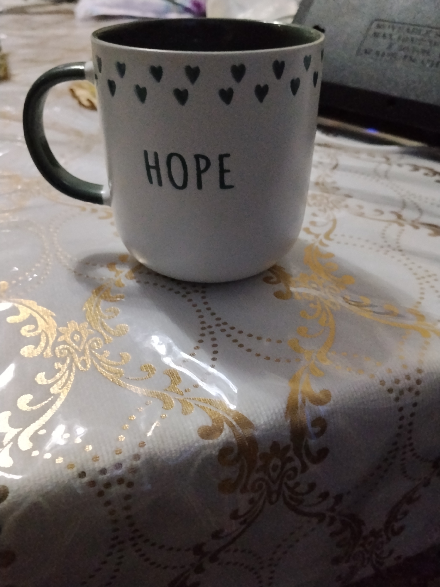 The image shows a close-up of a white ceramic mug with a dark green rim and handle. The mug is decorated with small, dark green hearts arranged in a pattern around the top, and the word "HOPE" is printed in black, sans-serif font in the center. The mug is placed on a table covered with a white tablecloth patterned with gold ornate swirls and small gold dots. The lighting is dim, casting a shadow of the mug on the tablecloth. There is a portion of a black electronic device visible in the upper right background, possibly a laptop or a monitor. The scene is indoors. There is no indication of the city of Bukhara, Uzbekistan, or any specific geographic location within it.
