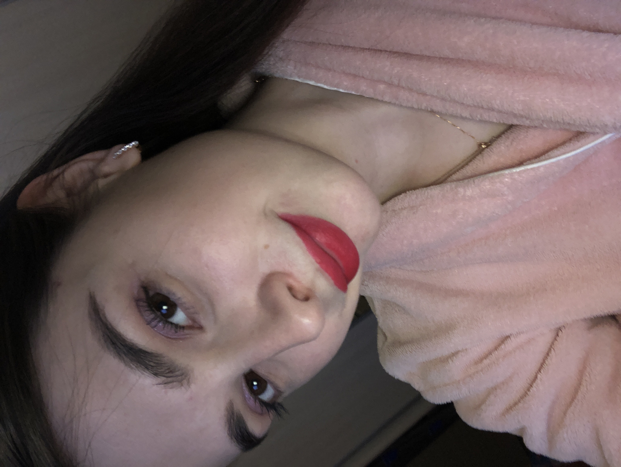 A close-up portrait features a young woman posing in what appears to be an indoor setting. The image is captured from a diagonal angle, focusing on her head and upper torso. She has long, dark hair, dark brown eyes, and prominent, well-groomed eyebrows. Her makeup includes bright red lipstick and a subtle, purplish hue applied around her lower lash line.
She is wearing a soft, light pink, fluffy robe or blanket, which covers her shoulder and chest. A delicate gold chain necklace with a small gold bead pendant is visible against her skin, partially obscured by the robe's opening. On her left ear, a small, sparkly ear cuff or earring adds a touch of adornment. The woman's gaze is directed towards the viewer, and her expression appears calm and neutral. The background is indistinct and dark, consistent with an indoor environment. The lighting suggests an indoor setting, and no specific time of day is discernible. There is no visible text in the image.