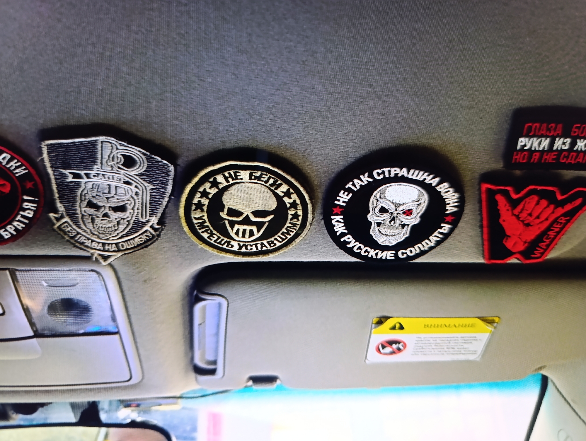 This image shows the interior of a vehicle, specifically the section above the front windshield. Affixed to the ceiling fabric are several patches or emblems, mostly featuring skull motifs. The patches include text in Russian, with visible words such as "CATER", "WAGNER", and phrases like "No right to be wrong" and "Not as scary as the Russian soldier". The patches vary in shape and color, with the Wagner patch being red and black. The sun visor is visible and flipped up, revealing a warning sticker in yellow with Russian text. The interior lights and rearview mirror are also visible. The setting appears to be inside a car, suggesting a personal space. The image does not provide enough information to identify it as Зипуново, Russia.