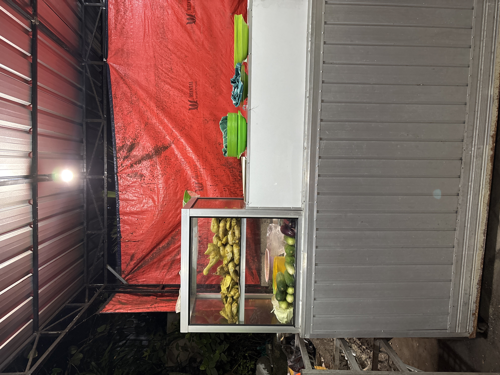 The image shows a food stall at night in Kuta Selatan, Indonesia. The stall has a silver metal base and a glass display case filled with fried food items on the top shelf and various vegetables, including cucumbers, cabbage, and eggplant, on the bottom shelf. In front of the stall, there are several bright green plastic bowls stacked, and two towels draped over them. The stall is covered by a corrugated metal roof supported by a metal frame. A large red tarp hangs behind the stall, with visible logos of "ORIENTAL." The scene is illuminated by a single light bulb hanging from the roof. The overall environment suggests an outdoor food vendor setup, typical of street food establishments in the region.