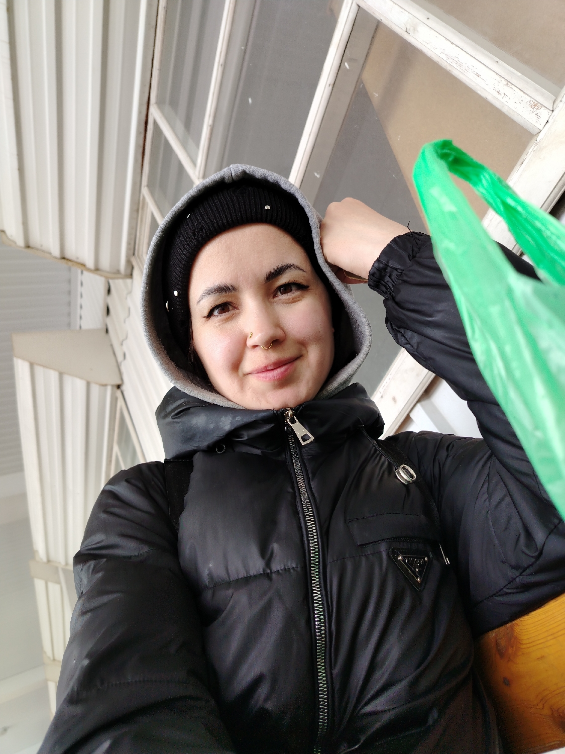 A close-up, low-angle shot captures a person, likely female, from the chest up. She has medium-dark skin, dark eyes with winged eyeliner, and a nose ring. She wears a black puffer jacket with a subtle sheen, a grey hooded sweatshirt with the hood pulled up, and a black beanie underneath, adorned with small embellishments. A small, triangular black patch with the word "PRADA" in silver text is visible on the upper right side of her jacket. She looks directly at the camera with a gentle, pleasant smile. Her left hand is raised, resting near her head, while her right hand holds a bright green plastic bag, partially visible on the right side of the frame.
The background features white corrugated metal siding on the left and top, suggesting a building or covered structure. A window with a white frame and what appears to be a screen is visible behind her on the right. A wooden bench is partially visible in the lower right. The subject's warm clothing, including a puffer jacket and beanie, indicates cool or cold weather, consistent with the location context of Chaikovsky, Russia. The lighting is soft and even, suggesting daytime, possibly overcast. The person appears to be waiting or resting.