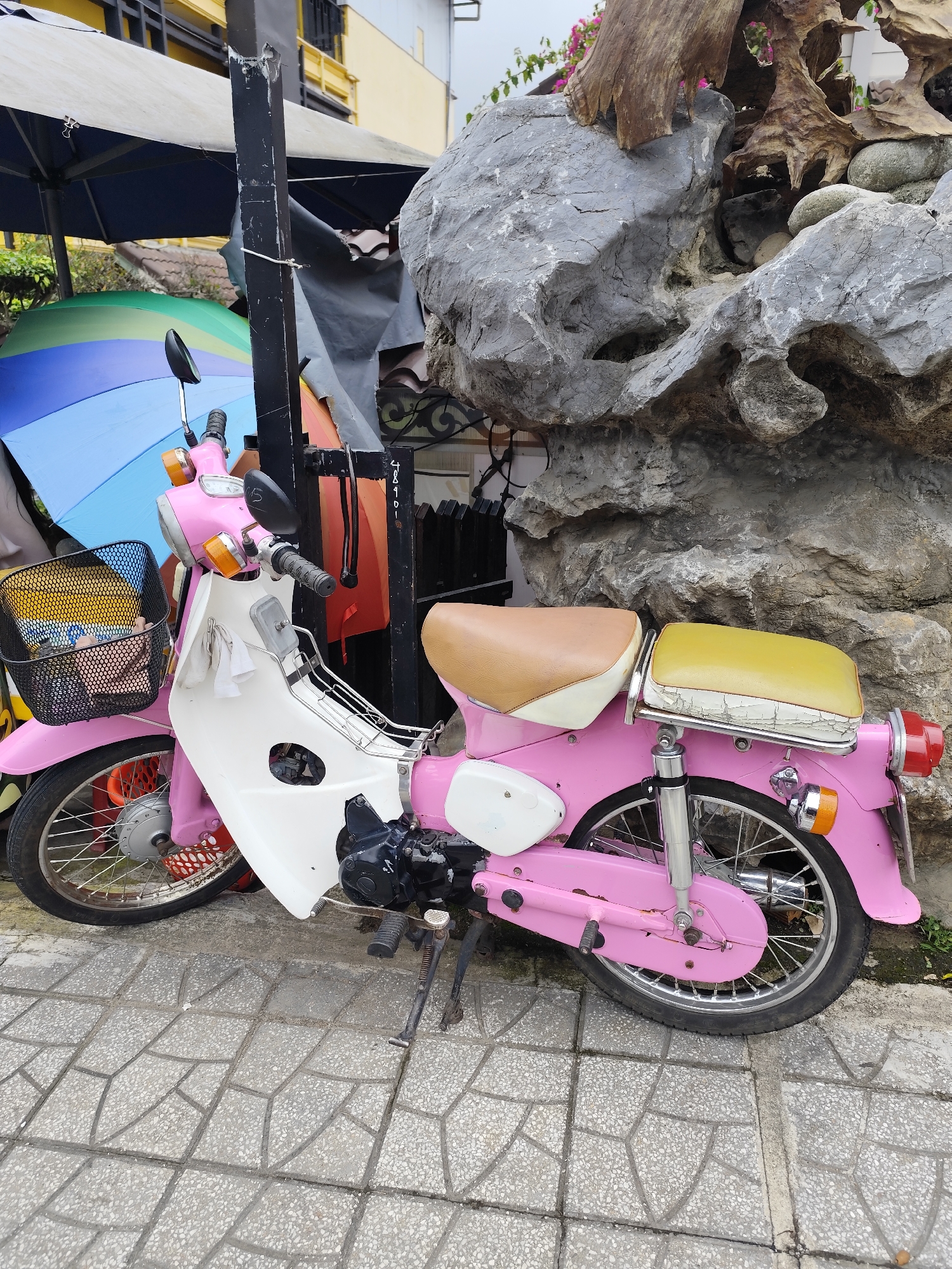 A pink and white moped is parked on a gray tiled sidewalk in Da Lat, Vietnam. The moped has a brown seat and a yellow rear passenger seat. A black wire basket is attached to the handlebars, containing a yellow object and a pink item. Behind the moped, a large, grey, textured rock formation is visible, with some driftwood incorporated into its structure. To the left of the moped, a colorful rainbow umbrella is partially visible, along with a blue umbrella. In the background, a yellow building with windows can be seen. A black metal pole stands next to the moped, with a white cloth hanging from it. The environment appears to be outdoors, possibly in a cafe or shop area.