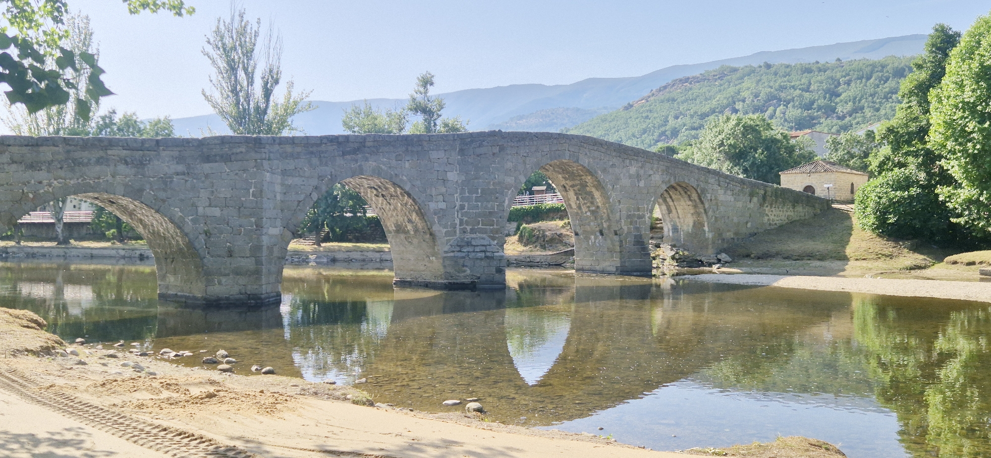 An arched stone bridge spans a calm river with sandy banks. The bridge, constructed from large, irregular stones, has multiple arches and is in good condition. Trees line the riverbanks, and a small, tan building with a tiled roof is visible on the right side of the frame, situated on a grassy incline behind the bridge. The background features lush green hills and mountains under a clear blue sky. The water reflects the bridge and surrounding greenery. Tire tracks are visible in the sand on the left bank.