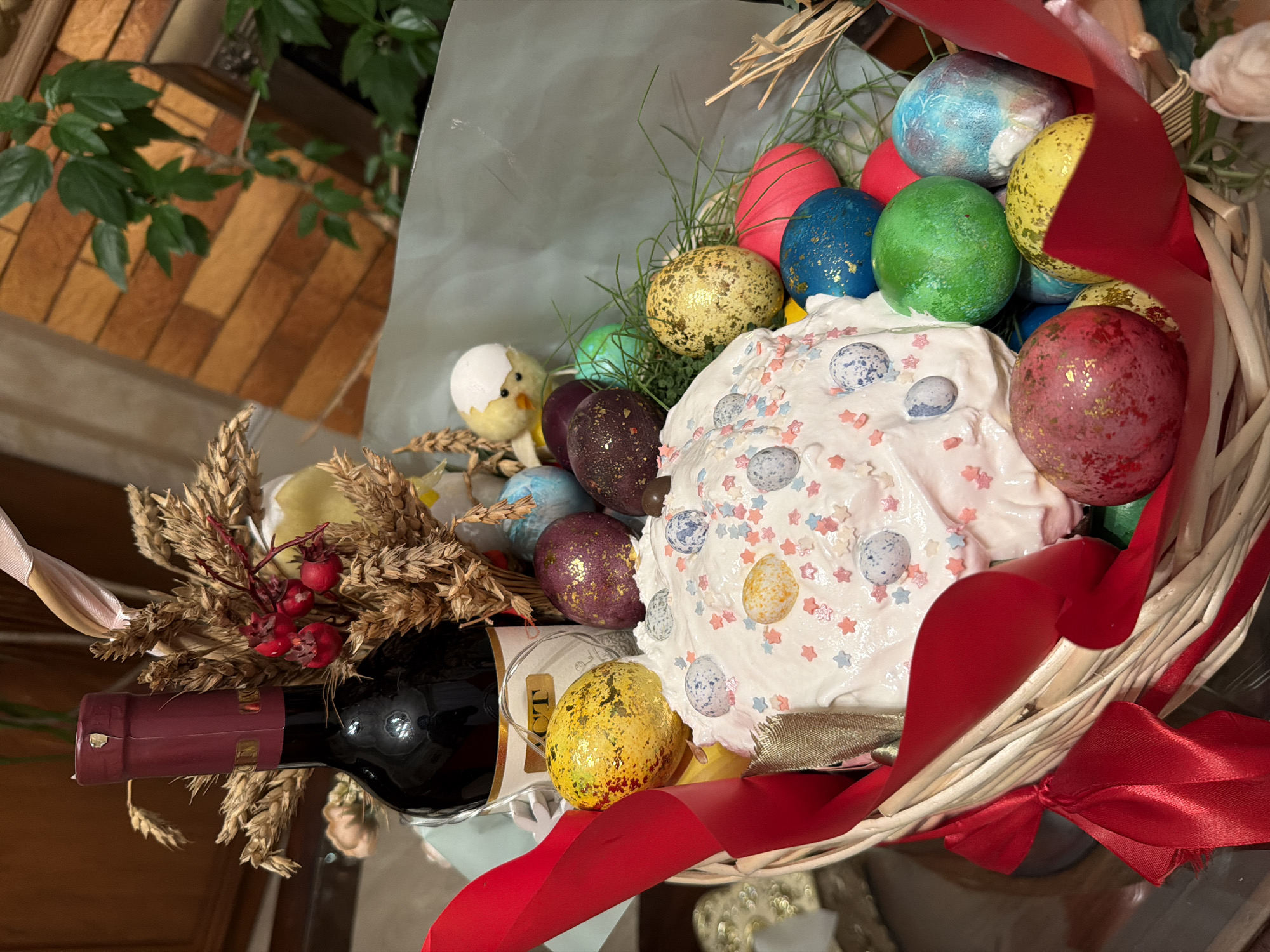A wicker basket filled with Easter treats is displayed indoors. The basket contains a tall Easter cake decorated with white icing and colorful sprinkles, several dyed Easter eggs in various colors (red, blue, green, yellow, purple, and a mix of gold), wheat stalks, a small plush chick, and a bottle of red wine. A red ribbon is tied around the basket. The background shows a glimpse of indoor vegetation and an orange and brown tiled wall, suggesting a room setting. It is not possible to determine the location in the city of Krasnoyarsk, Russia, from this image.