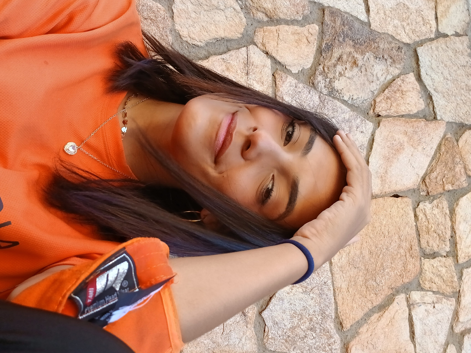 A woman with long, dark hair that transitions to blue at the ends is lying against a stone wall. She is wearing an orange long-sleeved shirt with a patch on the left shoulder. She has a gold necklace with a pendant. Her left hand is raised towards her face, with a blue bracelet on her wrist. The location appears to be outdoors, with the stone wall suggesting a possible residential or public space. No specific information suggests it is in Juan Griego, Venezuela.