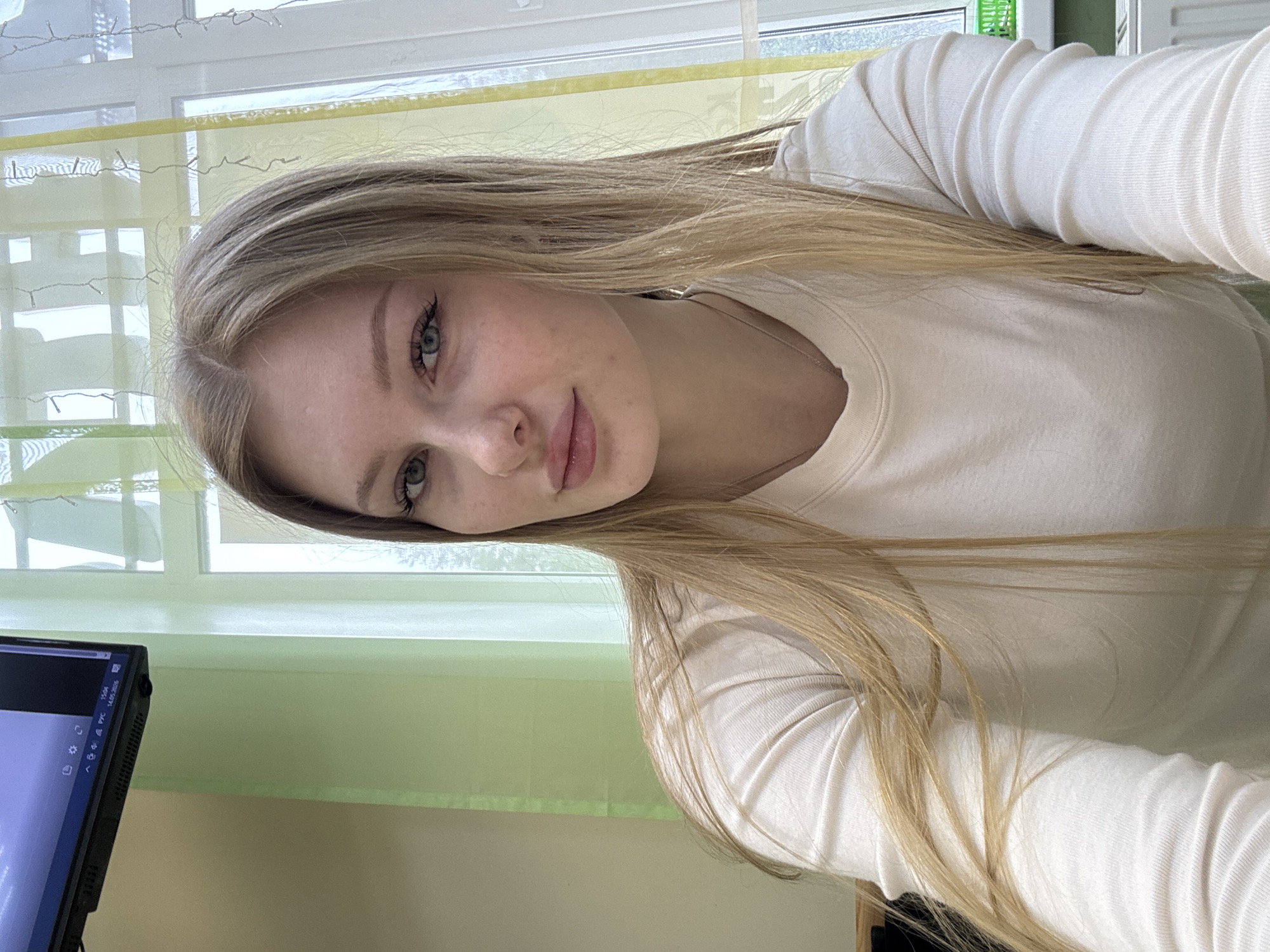 A young woman with light blonde hair and fair skin is posing for a selfie. She is wearing a light-colored long-sleeved shirt. Her hair is long and straight, falling over her shoulder. The background includes a window with sheer, light green curtains and a computer monitor on the left side of the frame. The location is Perm, Russia.