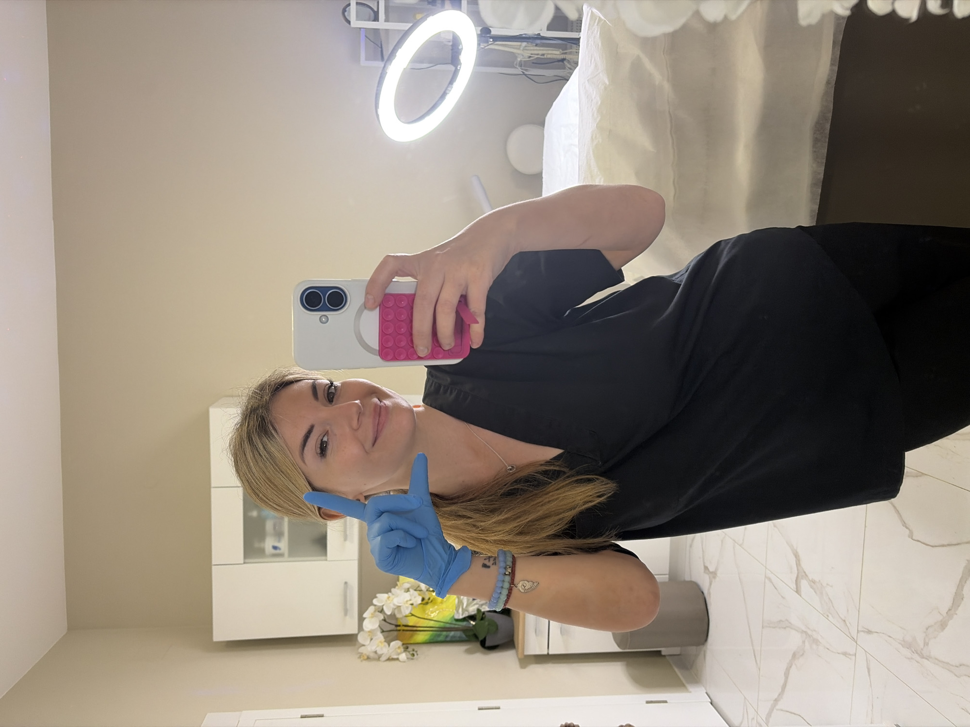 The image is a mirror selfie taken by a woman in what appears to be a beauty salon, spa, or aesthetic clinic located in Budva, Montenegro.
The primary subject is a fair-skinned woman with blonde hair, visible from the waist up. She wears a dark, possibly black, uniform-style top and a blue nitrile glove on her left hand. Her right hand holds a white smartphone, which has a prominent pink pop socket, indicating she is taking the mirror selfie. Her right wrist features several beaded bracelets.
The indoor setting has light-colored walls and a white marble-patterned tiled floor. In the background, white cabinets are visible to the left, with decorative white flowers placed below them. To the right, a white treatment bed covered with a light-colored sheet is partially in view. A bright ring light is positioned above and behind the woman, providing significant illumination.
The woman is smiling faintly and looking directly at her phone's camera in the mirror. Her gloved left hand is raised, forming a "peace sign" or "victory sign," conveying a cheerful and confident demeanor. The presence of the ring light, treatment bed, and medical-style glove strongly suggests a professional beauty or wellness environment where hygienic practices are observed. No discernible text is visible.
