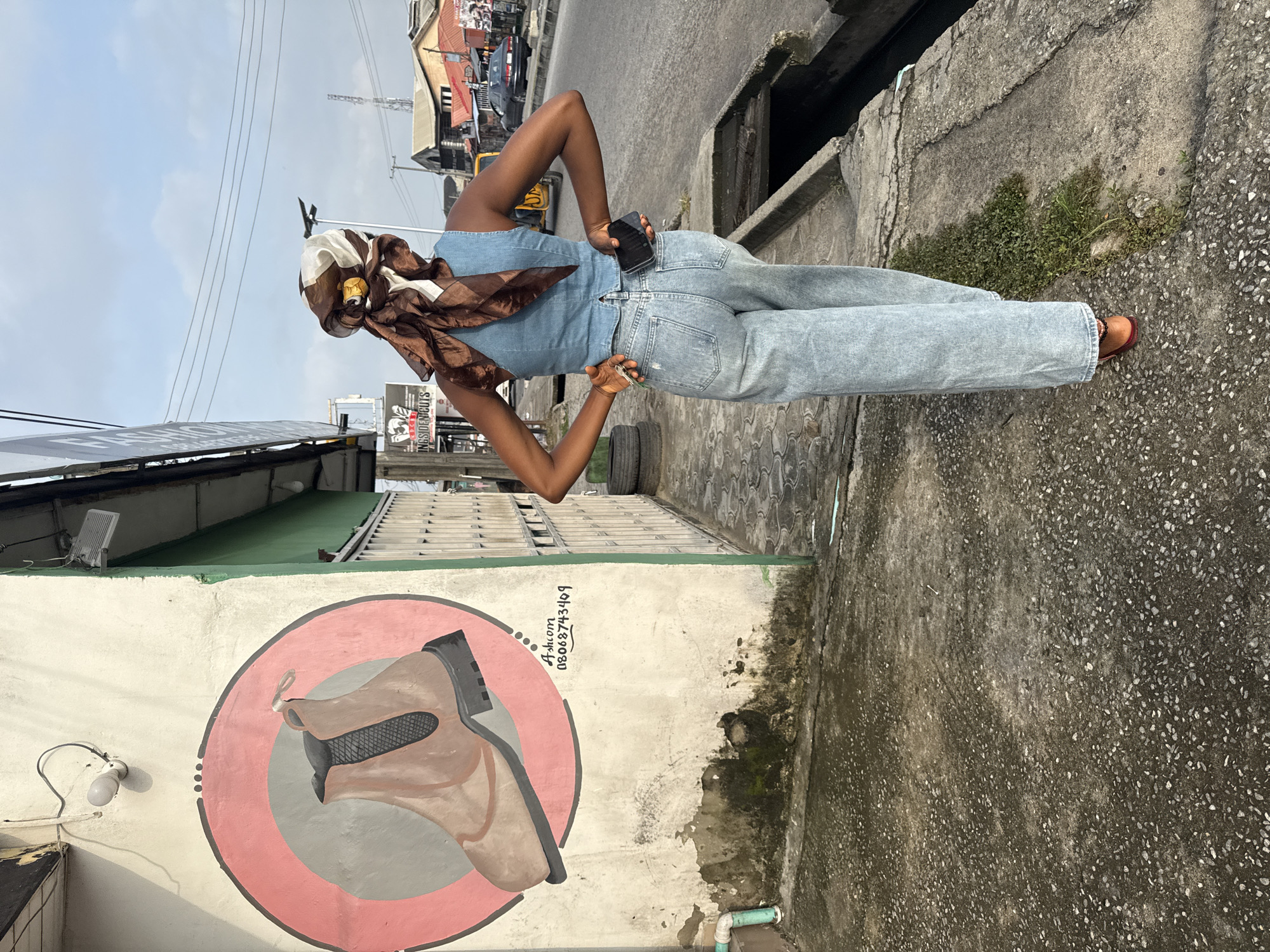 The image depicts a street scene in Ado, Nigeria. A woman, viewed from behind, stands on a sidewalk next to a wall adorned with a mural of a boot inside a circle. She wears a blue denim top, light blue wide-leg jeans, and an orange shoe. Her head is covered with a patterned headscarf in brown and white. She holds a phone in her right hand and keys in her left. The sidewalk is paved with concrete and has a drainage ditch running alongside it. In the background, there are buildings, a yellow motorized tricycle, and a black car on the street. The overall environment suggests a bustling urban setting.