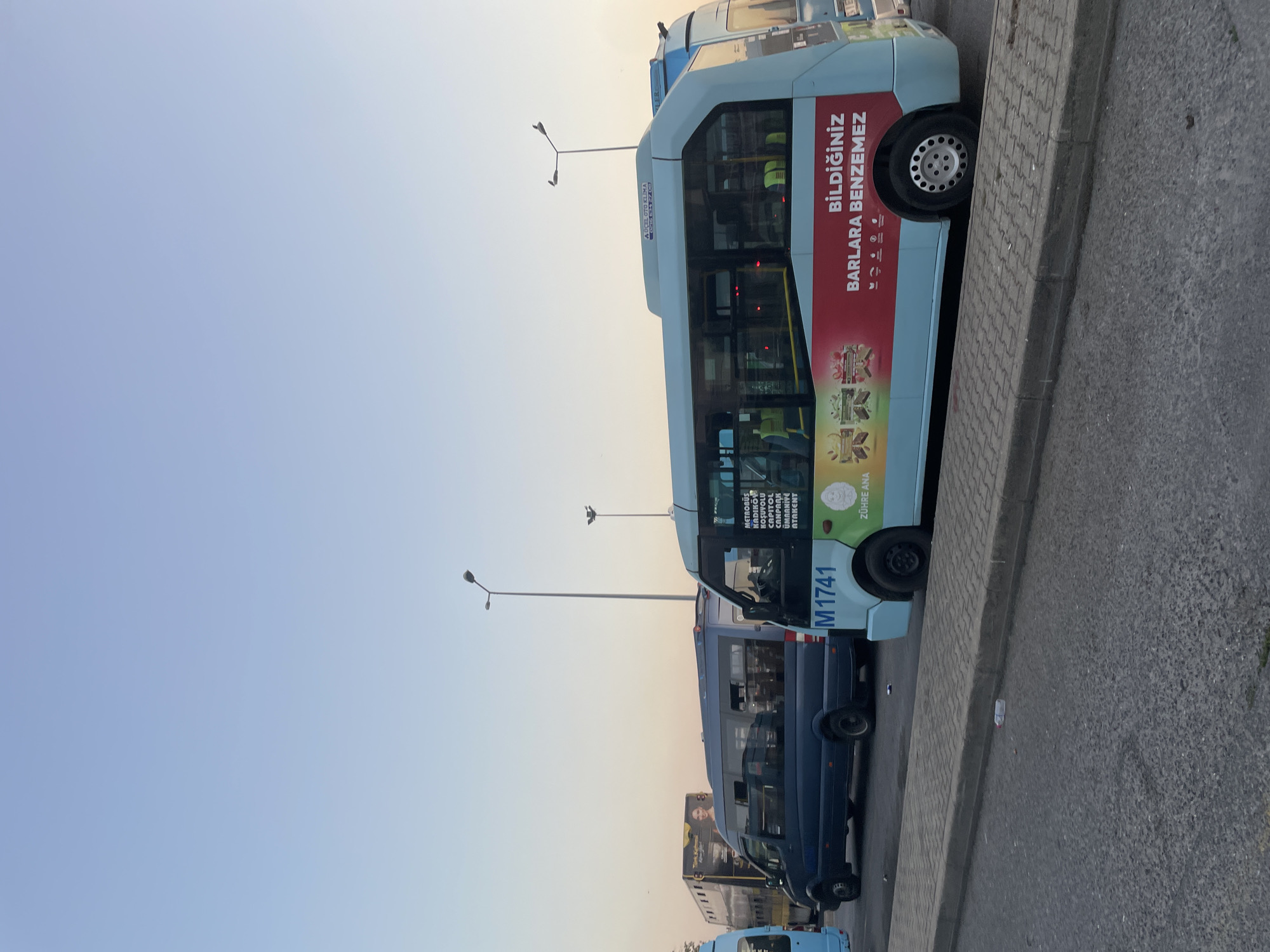 The image captures a street scene in Kadıköy, Turkey, likely a bus station or a busy road. In the foreground, a light blue bus with an advertisement on its side is parked. The advertisement features Turkish text, including "BİLDİĞİNİZ BARLARA BENZEMEZ," which translates to "It's not like the bars you know." Below this, a smaller text reads "ZÜHRE ANA." The bus's license plate is partially visible as "M1741."
Behind the light blue bus, a dark blue van is parked, appearing to be another form of public transport. Further in the background, a double-decker bus with an advertisement featuring a woman's face is visible. The overall setting suggests a bustling urban environment with multiple buses lined up. The sky above is a clear, pale blue, indicating it might be late afternoon or early morning. Streetlights are visible, one of which has two birds perched on it. The ground is paved with asphalt and a concrete curb separates it from a sidewalk made of interlocking pavers. A discarded plastic bottle lies on the sidewalk.