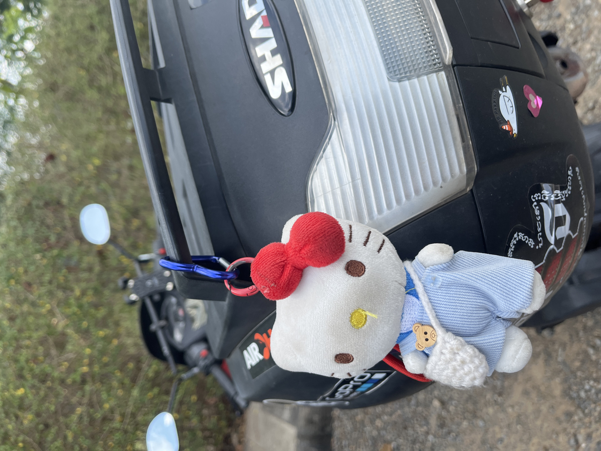 A close-up shot, oriented horizontally, shows a black Shad brand motorcycle storage case with a Hello Kitty plush keychain attached. The storage case is mounted on a motorcycle, which is partially visible with the background consisting of vegetation, suggesting an outdoor setting.
The black Shad storage case has the brand name "SHAD" prominently displayed in white letters. A clear plastic tail light is situated beside the brand name. Several stickers are affixed to the case, including one of a cartoon duck with a text that reads, "NO PARKING" and another sticker below that includes Cambodian script.
The Hello Kitty keychain is attached to the storage case with a red key ring and a blue carabiner. The plush toy is white with a red bow, brown eyes, a yellow nose, and is dressed in a light blue outfit with a small, white, knitted purse.
The motorcycle has visible handlebars, mirrors, and some of its body. The background vegetation includes grass and possibly some trees.
The image was taken in the city of Ban Tom, Thailand.