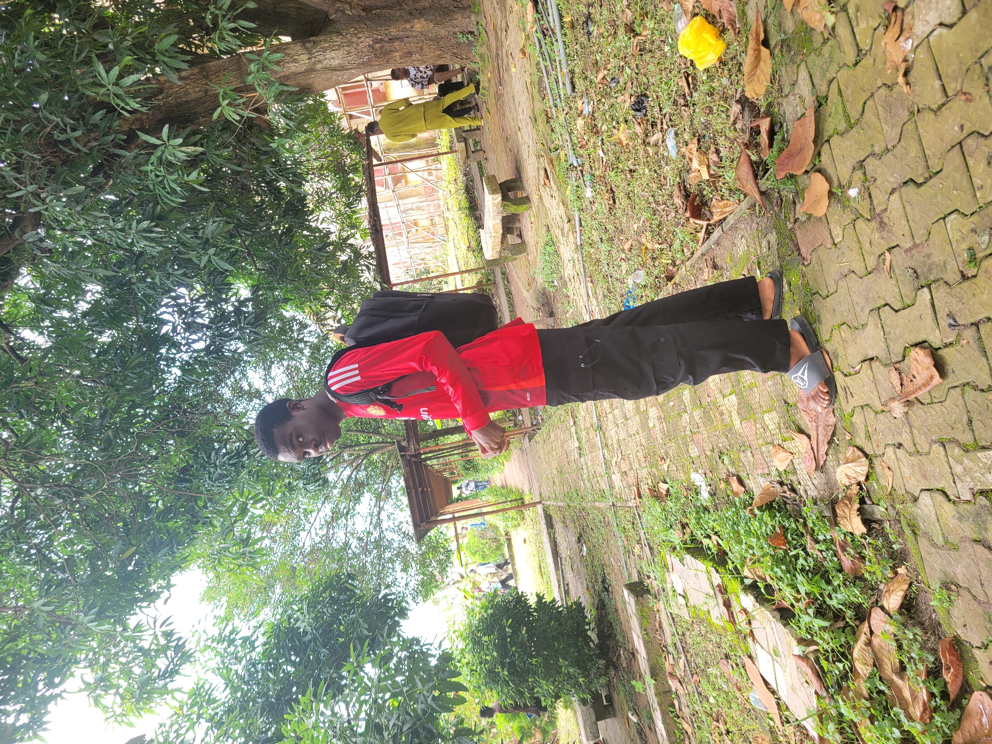 In Ukukwa, Nigeria, a young Black male stands on a brick pathway in a park-like setting, facing the camera. He is wearing a red long-sleeved shirt with white stripes on the sleeves, black cargo pants, and gray slides. A black backpack is slung over his right shoulder. The pathway is lined with green foliage and large trees. In the background, other individuals are visible: one person in a yellow outfit walks away from the camera, and another person sits on a stone bench next to a weathered structure. The ground is covered with leaves and some scattered litter.