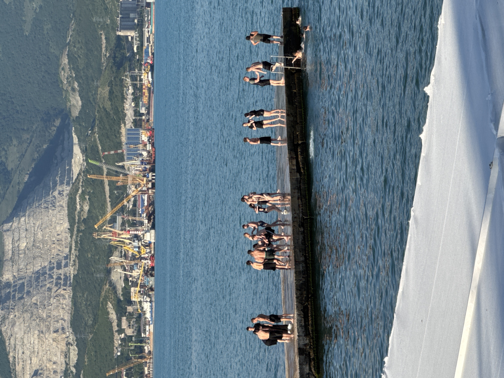 The image depicts a group of people on a concrete pier extending into a body of blue water. The pier is situated in front of a busy industrial port, characterized by numerous cranes, ships, and buildings. In the background, a large, terraced mountain dominates the skyline. Some individuals on the pier are dressed in swimwear, suggesting a recreational setting, while others are in casual clothing. Several people are in the water, swimming or preparing to swim, with some using a ladder to access or exit the pier. The overall atmosphere appears to be a sunny day at a coastal industrial area. This scene is consistent with locations in the port of Novorossiysk, Russia, known for its extensive industrial facilities and proximity to mountainous terrain.