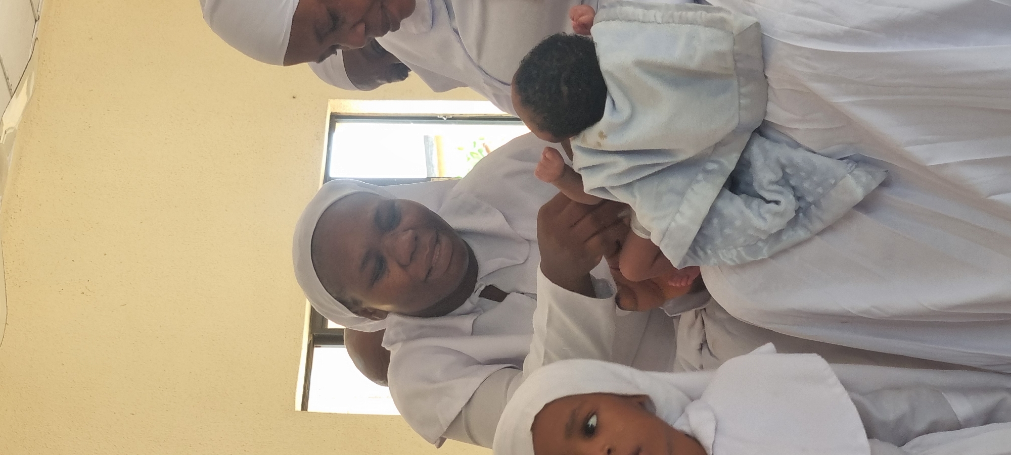 The image shows four individuals. Three adults, all wearing white robes and head coverings, are attending to a baby wrapped in a light-colored blanket. The woman in the foreground is looking down at the baby with a gentle expression. A child is also present, dressed in similar white attire. The scene appears to be indoors, with a window visible in the background. The specific location in Lagos, Nigeria, cannot be determined from the image.