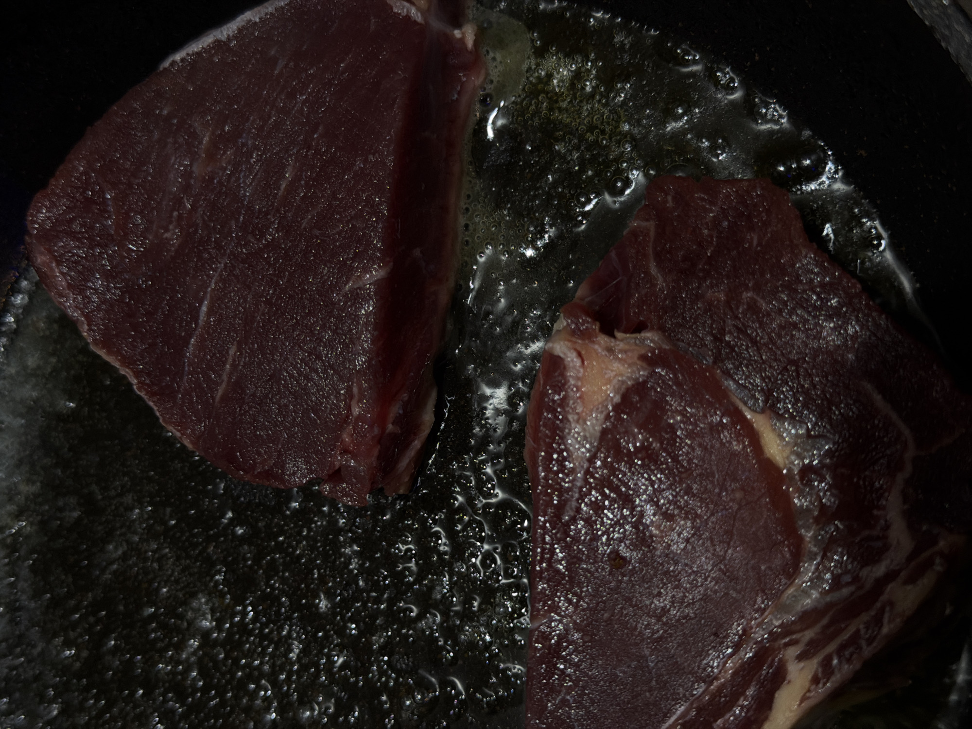 The image shows two pieces of raw meat, likely beef, cooking in a pan with oil or fat. Bubbles are visible in the oil. The scene appears to be indoors. The location, North Kuta, Indonesia, is extraneous information as there is no visual evidence in the image to support this.
